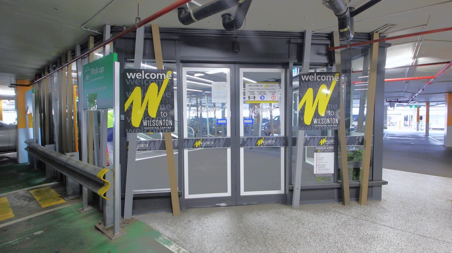 Entry to the Wilsonton Shopping Center from the undercover carpark is via automatic, glass sliding doors with grey metal frames. On both sides of the front of the door are two narrow wood-like metal frames placed diagonally holding the two black signs for ‘Welcome to Wilsonton Shopping Center’. The carpark has concrete flooring and ceiling with numerous pipes and mechanical features.