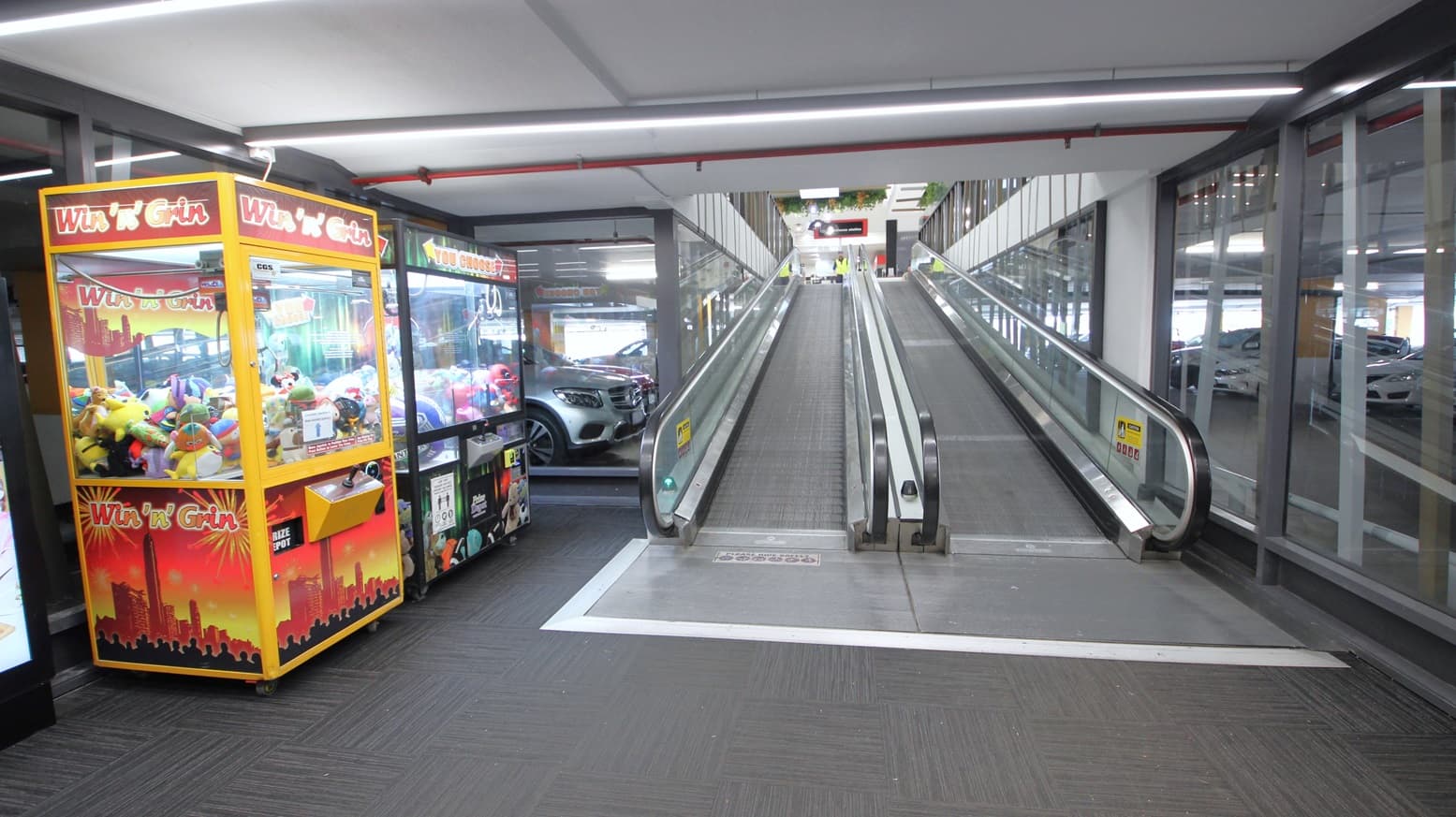 Two escalators leading up to the Wilsonton Shopping Center accessible from the undercover carpark. The entry section has grey carpet floors. On the left side are yellow and black toy machines. The small section before the escalators is painted in grey with a white outline. Seen through the glass walls are cars parked on the available 800 free car parking spaces.