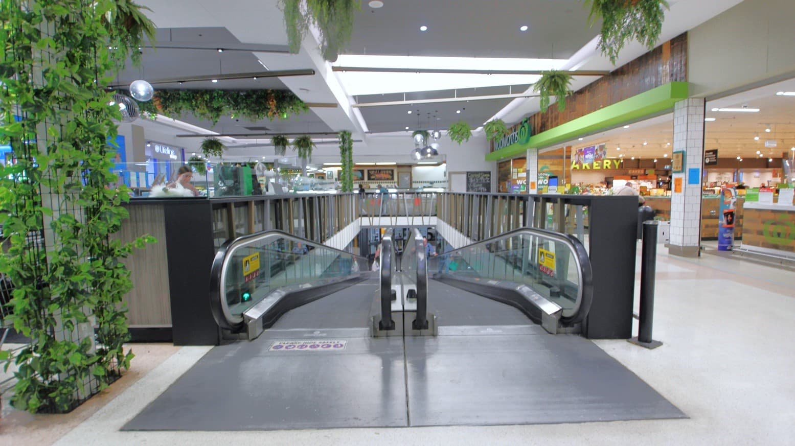 Two escalators from the undercover carpark of Bridge Street with 8000 free car parking spaces. On the left side is a column enclosed by a black screen that has green vines. Covering the other specialty retailer sections is a dark wooden handrail with light blue, brown, and grey diagonal walls. Hanging on the ceiling are green plant pots.