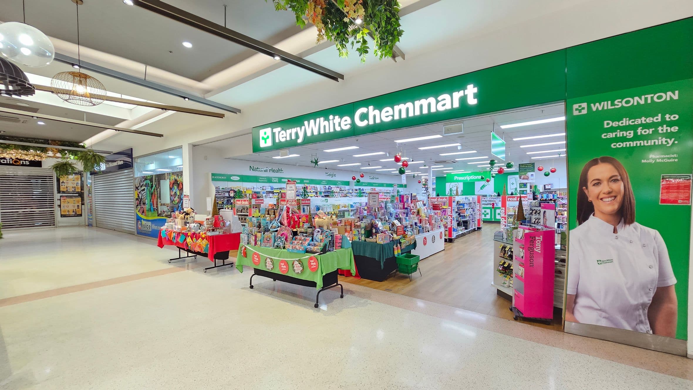 A pharmacy entrance with a bright green sign reading "TerryWhite Chemmart" above. To the right, a large poster features a smiling person in a white uniform. Inside, shelves are stocked with various products, and tables display items with festive decorations. The floor is light-coloured, and the ceiling has recessed lighting and hanging plants. On the left, a closed shop with a metal shutter and a colourful display in front.
