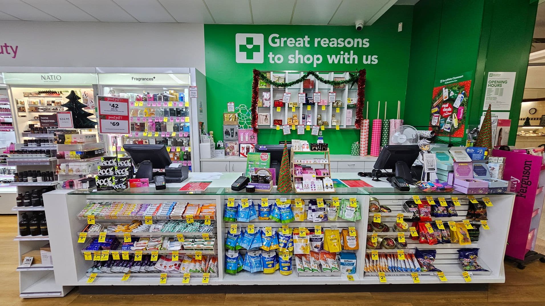 A pharmacy checkout counter with various products displayed. In the foreground, shelves are stocked with snacks and small items, each with yellow price tags. The counter has two registers and is decorated with holiday-themed items. Behind the counter, a green wall features a sign with a cross and promotional text. Shelves on the wall display more products, some with festive decorations. The floor is wooden, and the ceiling is white with recessed lighting.