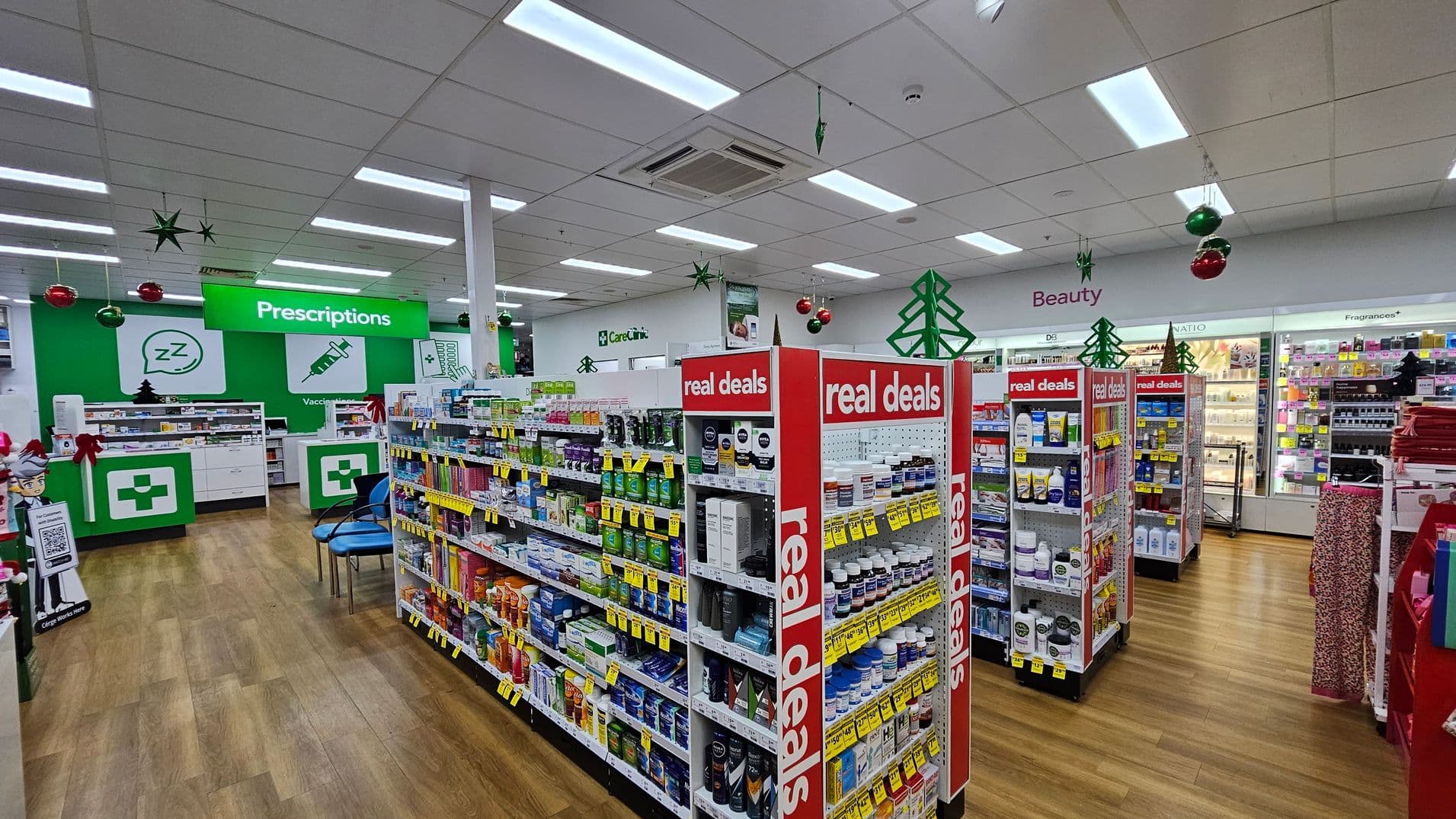 A pharmacy interior with wooden flooring and a white ceiling. On the left, there's a green "Prescriptions" sign above a counter with a cross symbol. Shelves with various products are in the centre, marked with "real deals" signs. On the right, a section labelled "Beauty" displays cosmetics. Christmas decorations, including green stars and red baubles, hang from the ceiling.