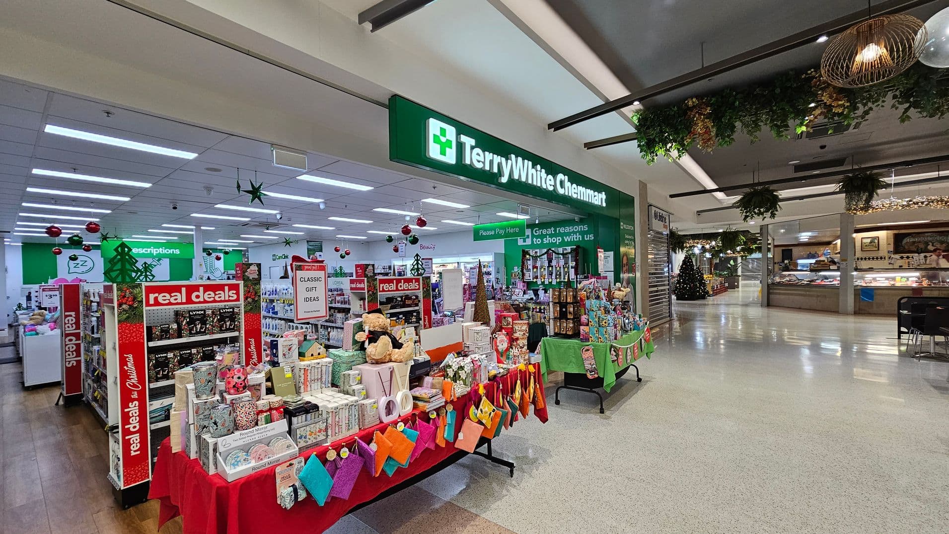 A pharmacy with a green sign reading "TerryWhite Chemmart" is on the left. Inside, there are displays with "real deals" signs and various gift items on red and green tablecloths. Christmas decorations, including baubles and stars, hang from the ceiling. On the right, there's a shopping centre area with a Christmas tree and food outlets. The floor is light-coloured, and the ceiling has hanging plants and lights.
