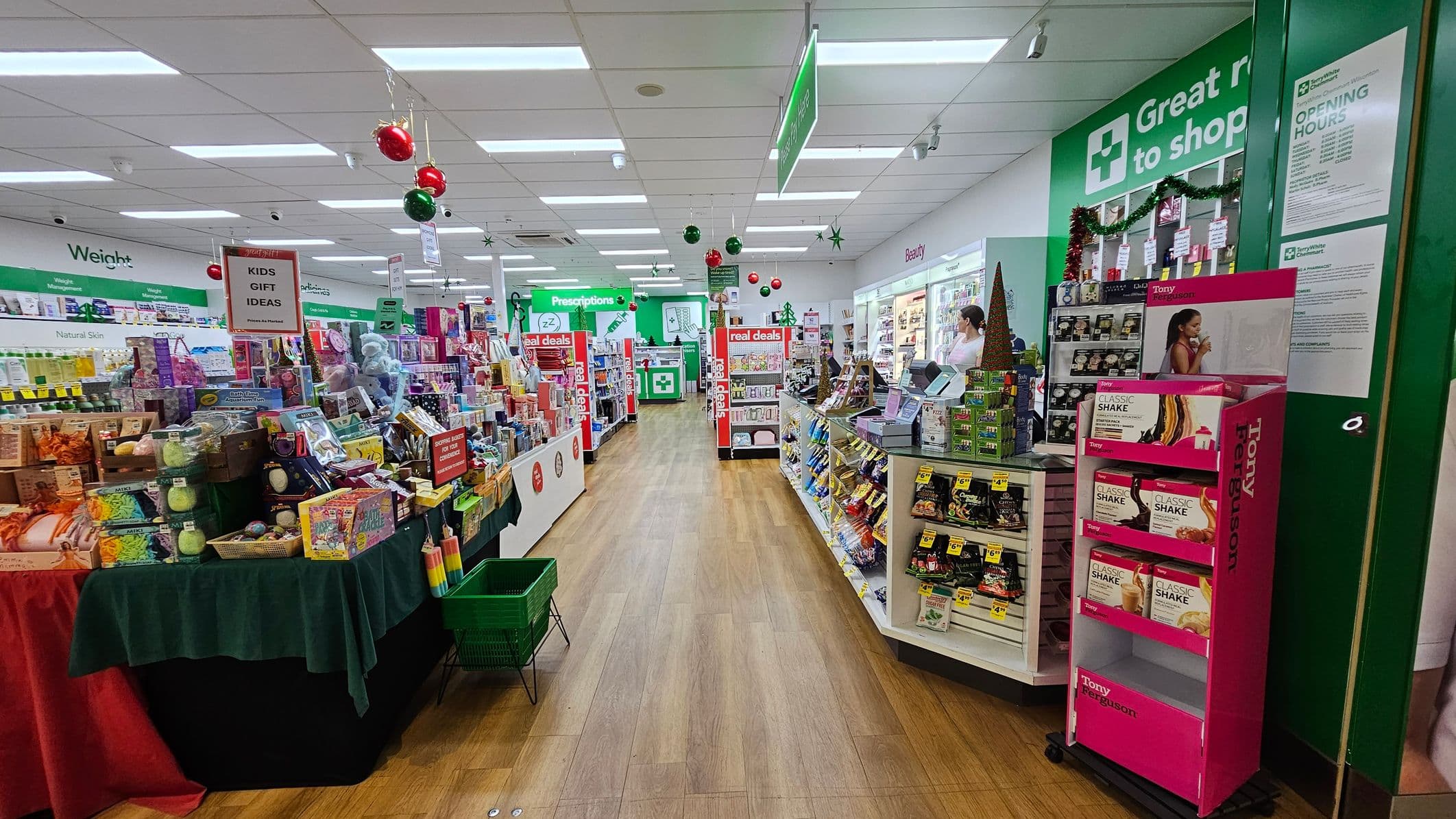 A pharmacy interior with a wooden floor and a white ceiling with fluorescent lights. On the left, there are shelves filled with various gift items under a sign reading "KIDS GIFT IDEAS." On the right, there are shelves with beauty products and a display for "Tony Ferguson" shakes. In the background, a counter with a green sign for prescriptions is visible. Red and green decorations hang from the ceiling, and a sign with opening hours is on the right wall.