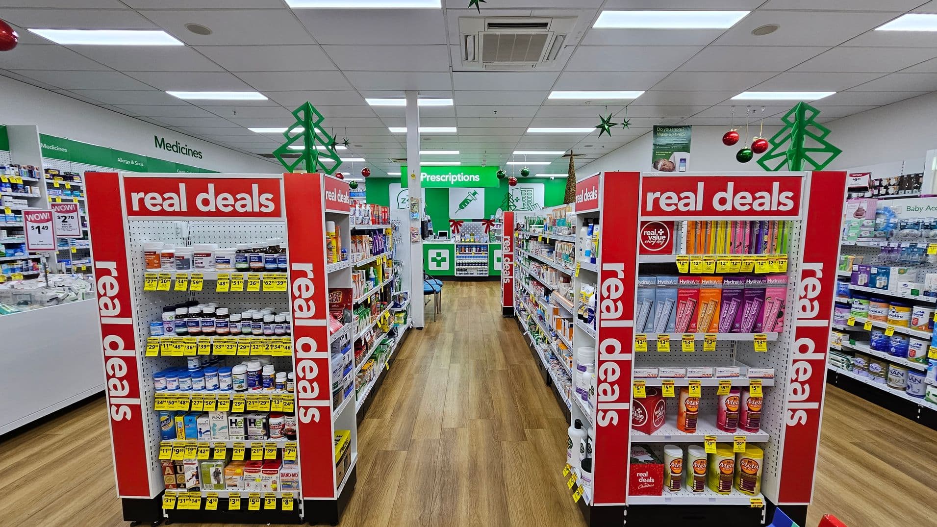 A pharmacy interior with two central aisles displaying products under "real deals" signage. The shelves are stocked with various health and wellness items, each with yellow price tags. The background features a green "Prescriptions" sign above a counter. Green star and red bauble decorations hang from the ceiling, with green tree shapes visible. The floor is wooden, and the ceiling is white with fluorescent lighting.