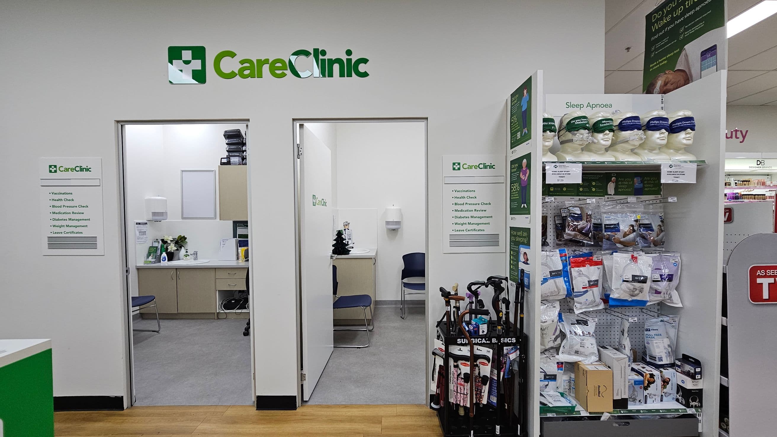 On the left, a CareClinic sign is above two open doors leading to consultation rooms with chairs and medical supplies. In the middle, a sign lists services like vaccinations and health checks. On the right, a display for sleep apnoea products features mannequin heads with masks, and shelves with related items. A stand with walking canes is nearby. The floor is a mix of wood and grey tiles, and the ceiling has fluorescent lighting.