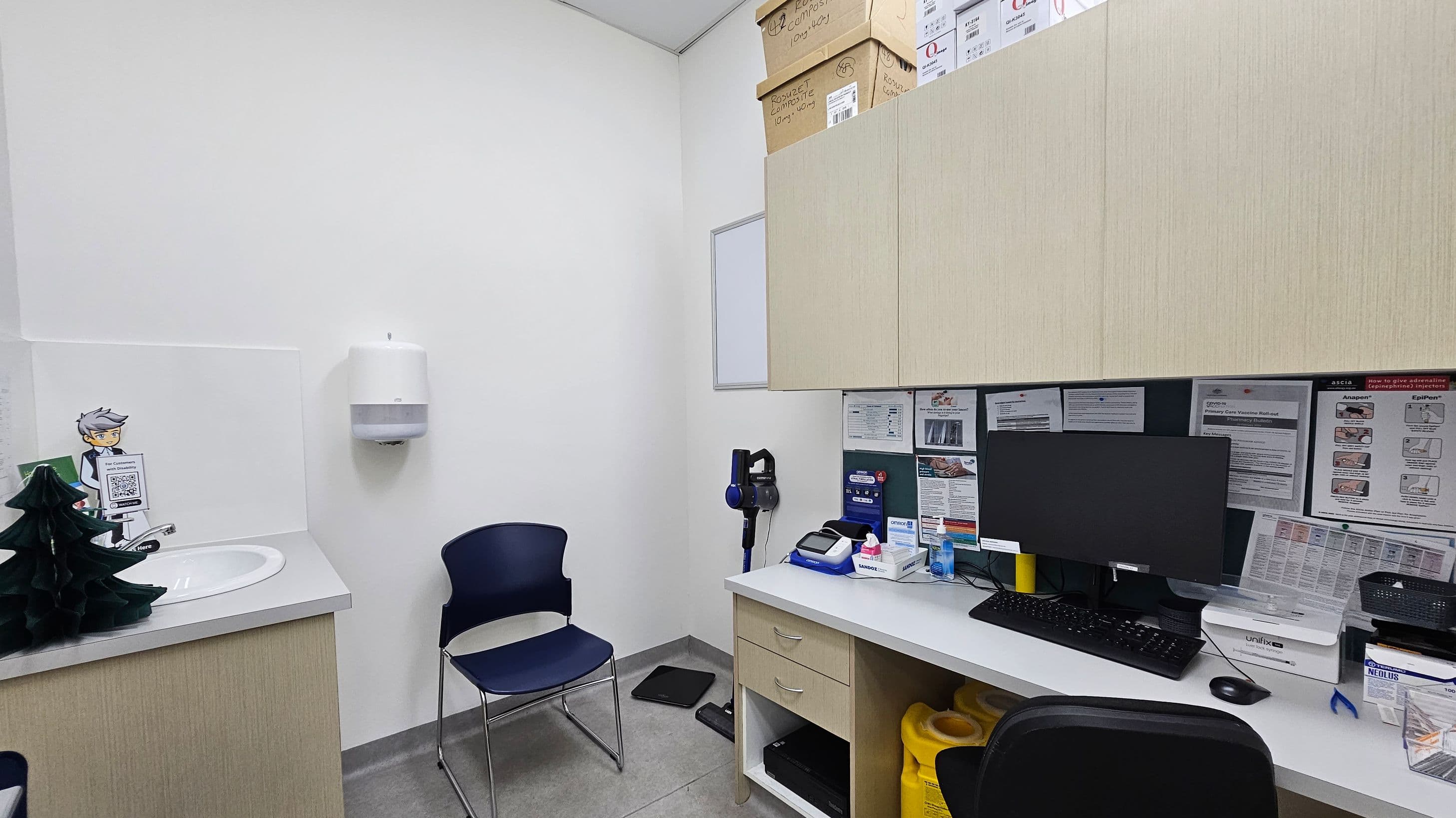 A small office or clinic room with a white wall and grey tiled floor. On the left, there's a sink with a paper towel dispenser above it and a small decorative tree. A blue chair is positioned against the wall. On the right, a desk with a computer monitor, keyboard, and various office supplies. Above the desk, there are light-coloured cabinets with boxes on top. The wall behind the desk is covered with notices and information sheets.