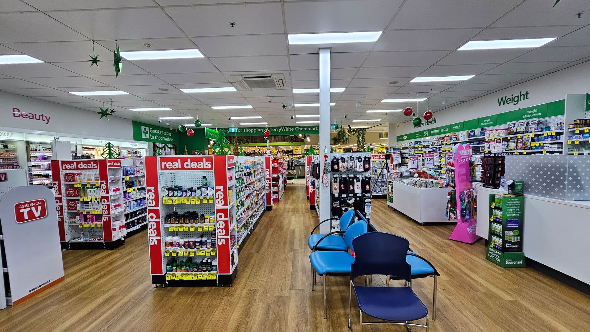 Inside a pharmacy with wooden flooring and a white ceiling. In the foreground, there are blue chairs on the right. The middle features aisles with shelves labelled "real deals" filled with various products. The left side has a section marked "Beauty" with cosmetics. The right side displays "Weight Management" products. Green and red decorations hang from the ceiling, and a sign in the background reads "Great reasons to shop with us."