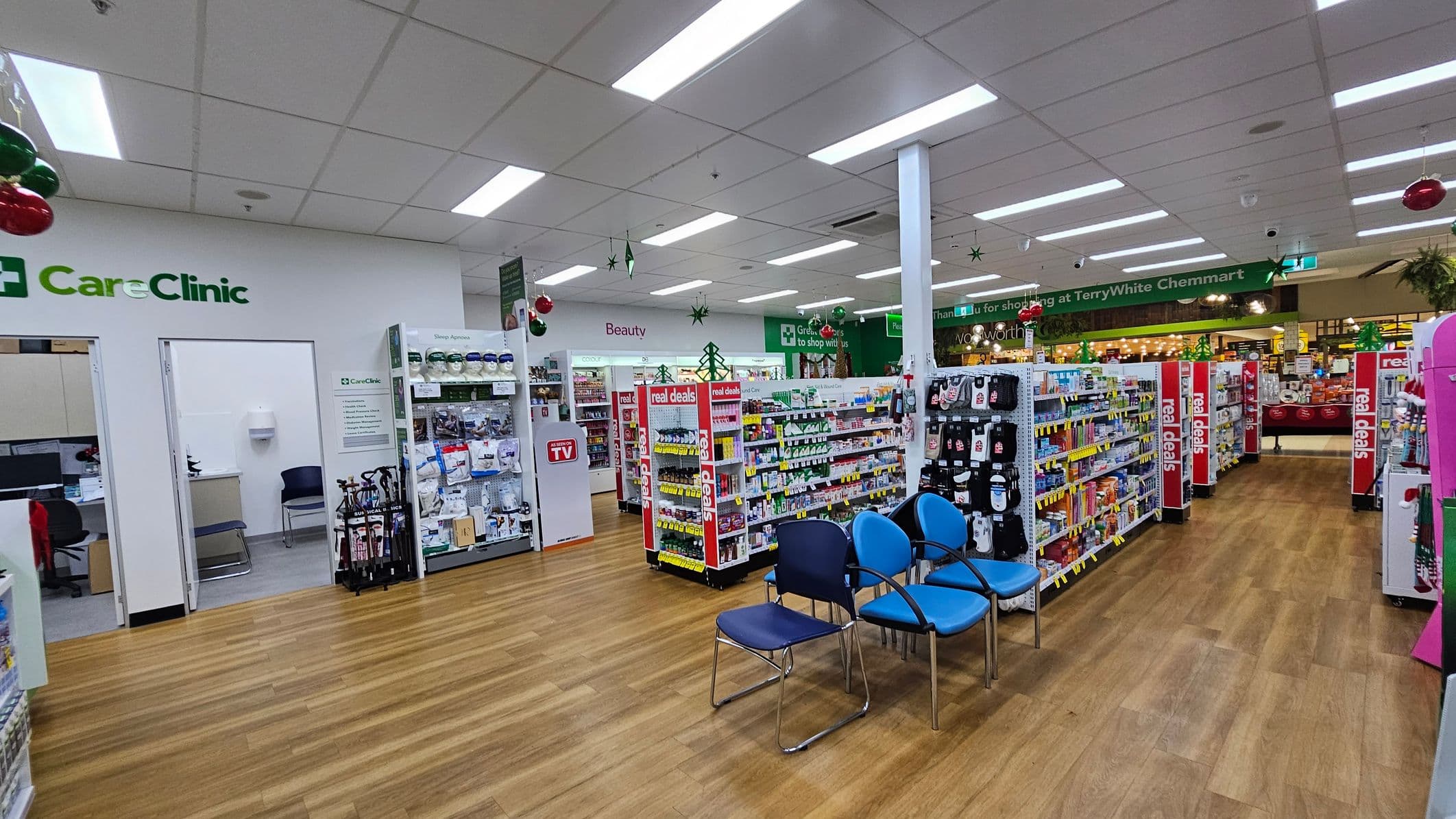 Inside a pharmacy with a wooden floor and a white ceiling with fluorescent lights. On the left, there's a "CareClinic" area with open doors leading to consultation rooms. In the centre, shelves display various products, including a section labelled "Beauty." Blue and black chairs are arranged in the foreground. On the right, more shelves with "real deals" signs. The background shows a green sign thanking customers for shopping, with a glimpse of another store area.