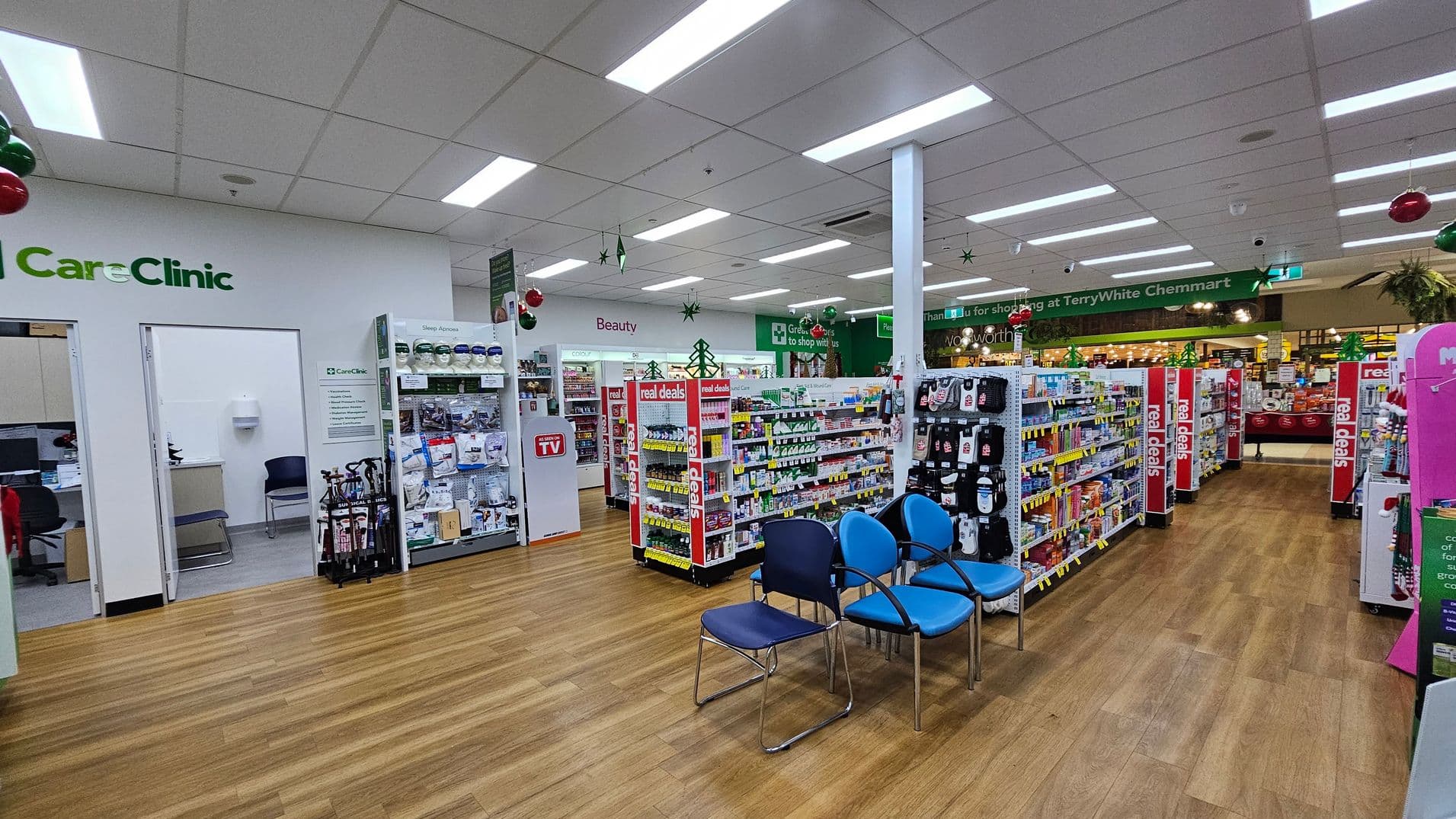 Inside a pharmacy with wooden flooring and a white ceiling with fluorescent lights. On the left, there is a "CareClinic" area with open doors leading to consultation rooms. In the centre, shelves display various products, with signs indicating "Beauty" and "real deals." On the right, there are blue chairs for seating. The background shows more shelves and a green sign thanking customers for shopping. Christmas decorations, like baubles and stars, hang from the ceiling.