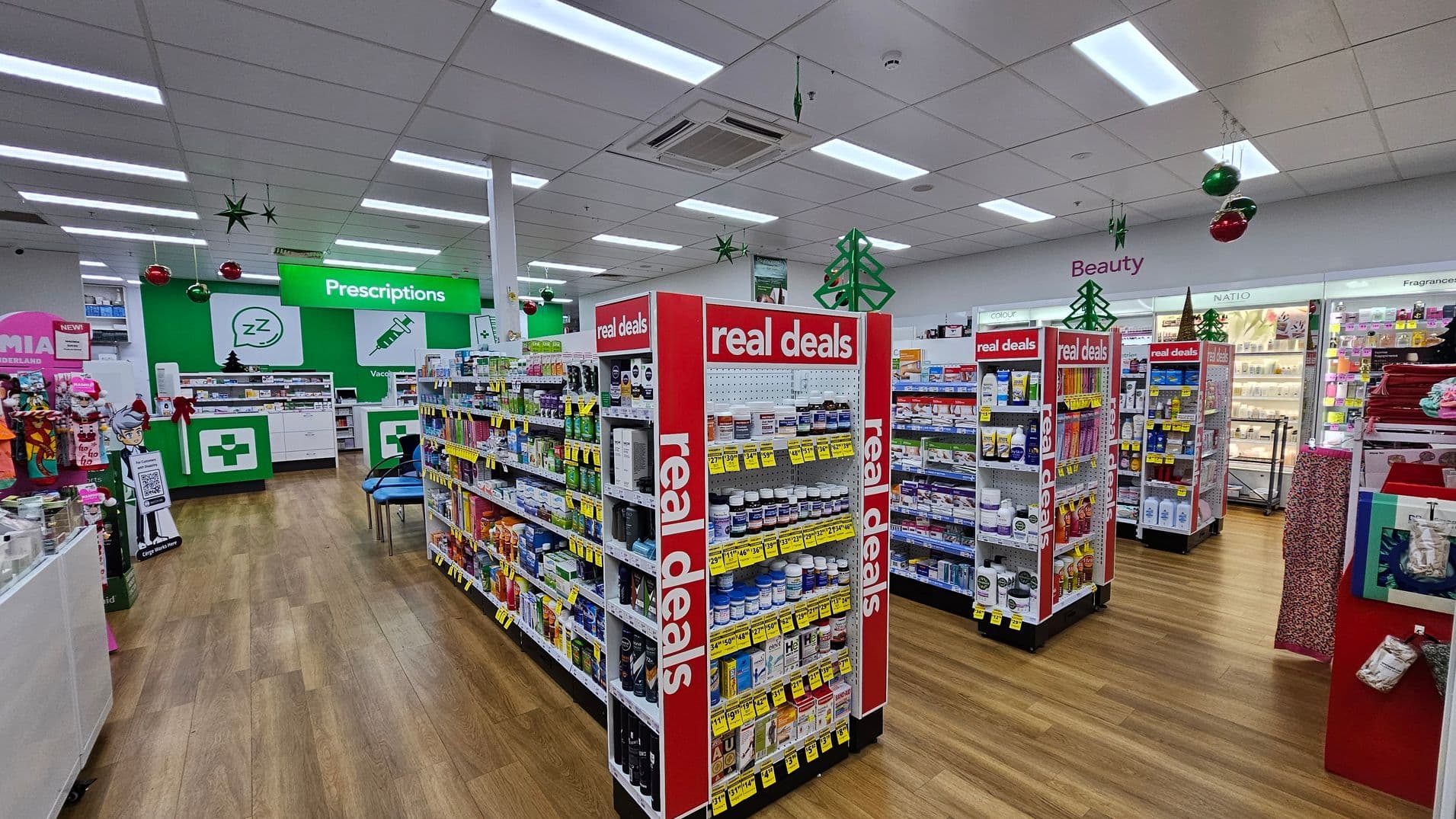 Pharmacy interior with wooden flooring and a white ceiling. In the centre, there are shelves labelled "real deals" displaying various products. To the left, a prescriptions counter with a green sign and festive decorations, including hanging stars and baubles. On the right, a beauty section with illuminated shelves showcasing cosmetics. Green and red Christmas decorations are visible throughout the store.