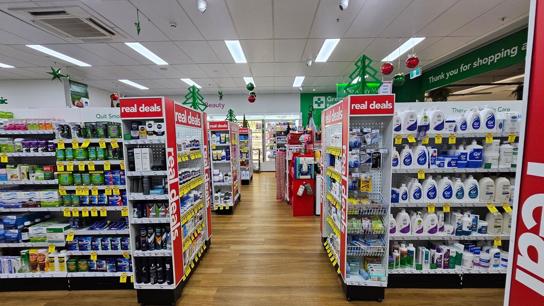 A pharmacy interior with wooden flooring and a white ceiling featuring fluorescent lights. In the foreground, two aisles display various health and beauty products, with red signs reading "real deals." The left aisle has smoking cessation products, while the right aisle displays skincare items. In the background, a refrigerated section is visible, and green and red decorations hang from the ceiling. A green sign above reads "Thank you for shopping."