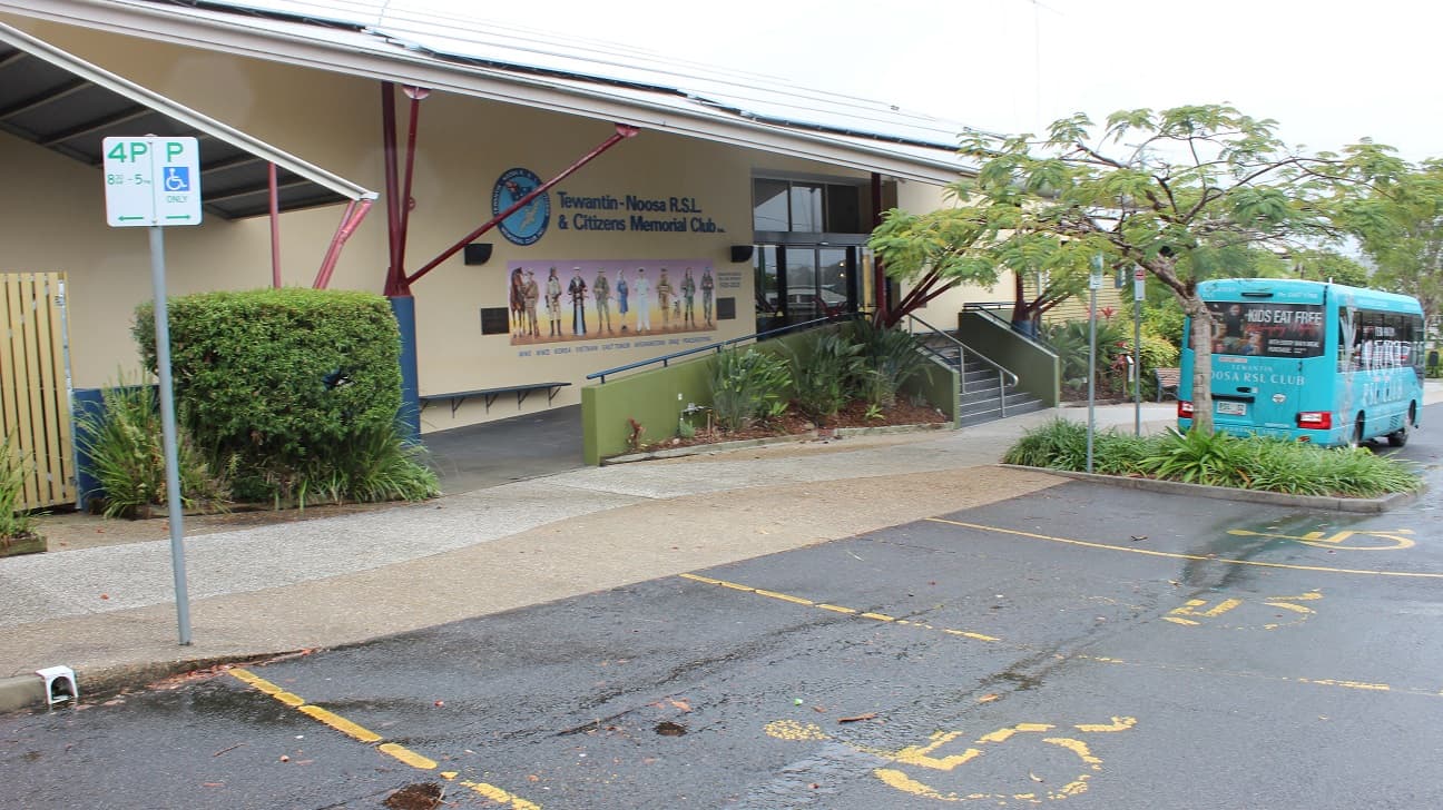 The front wall with military artwork, the Tewantin Noosa RSL badge, two etched tiles with inscriptions, and blue-painted seats on the yellow wall under a facade. The front door is accessed through a two-side ramp access walkway and a stepped walkway in the centre flanked by green-painted half-walls and steel railings facing a sidewalk and a parking lot. The wall is surrounded by sculpted areas of grass and a tree. Three direction signs are visible.
