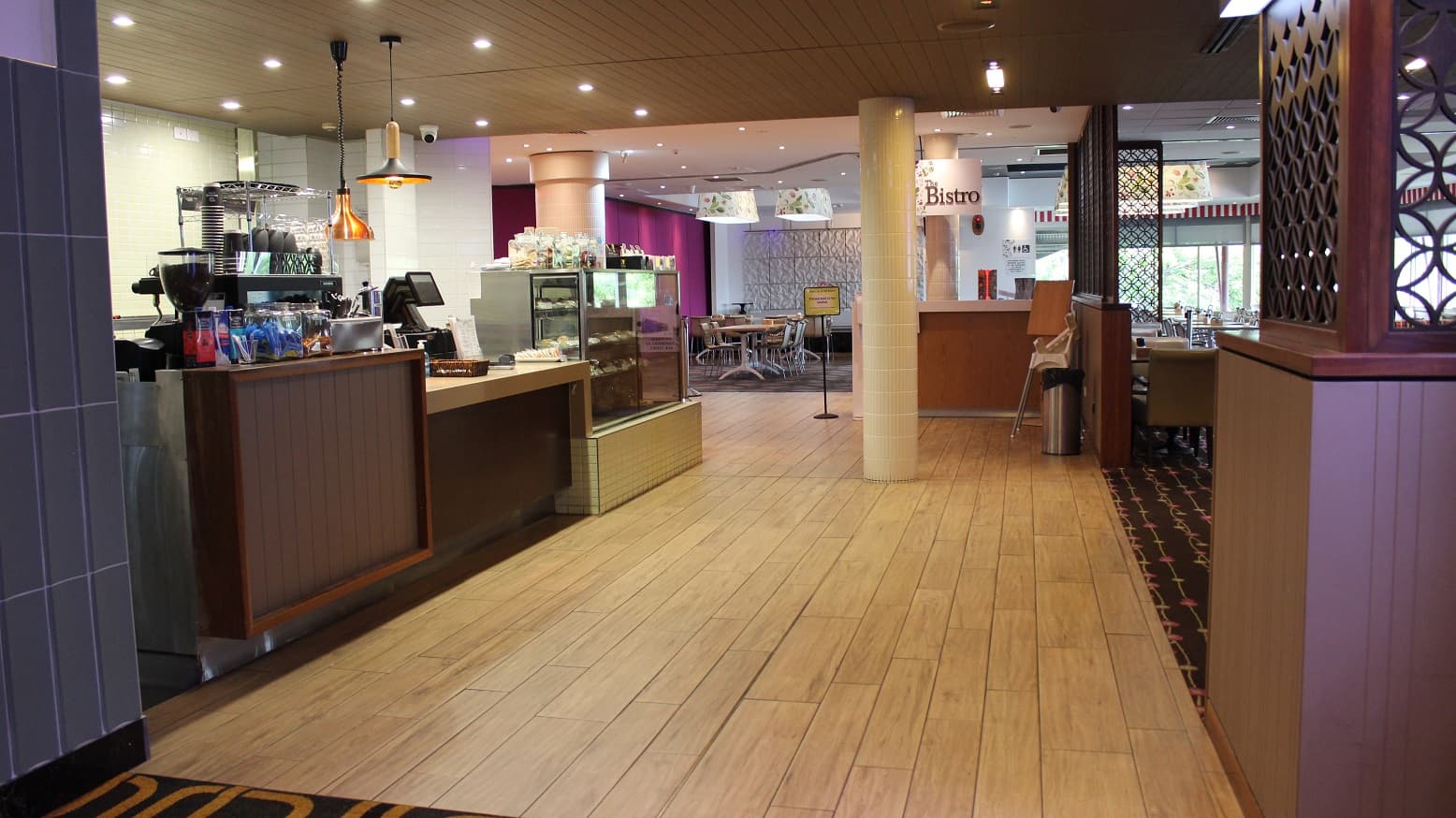 The Bistro restaurant is accessed through a beige-covered floor, with a brown designed counter on the left having coffee packs,a cabinet with cups, a checking screen, and a lit snacks showing cabinet. On the left is a wide entrance with walled space having a rubbish bin and seats. Two white pillars face the bistro tag on a flowering white lamp cover and the direction board on the entry to the purple and white wall dining room with seats and tables on the right.
