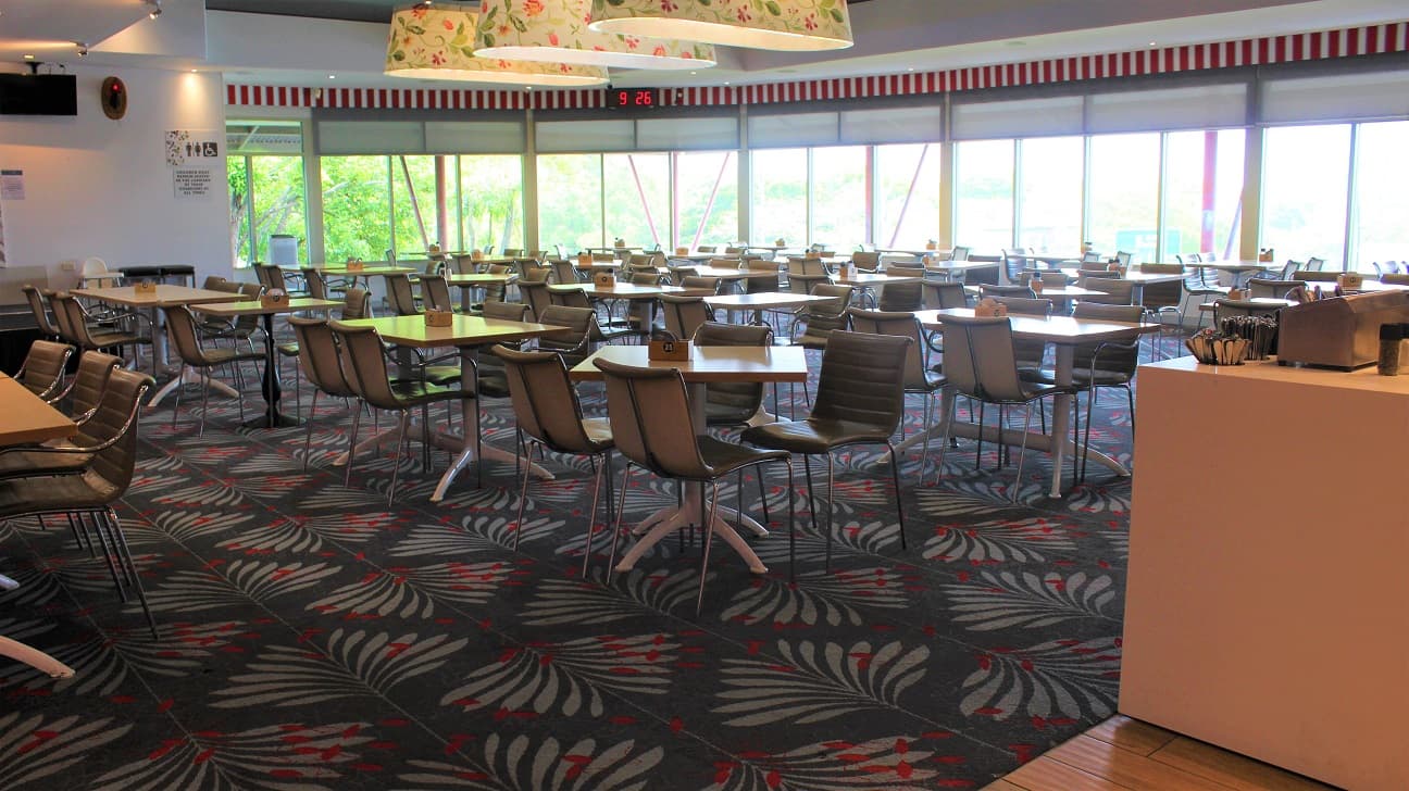 A dining hall at the Bistro restaurant covered with grey and crimson designed tiles. Having four seats to a table set vertically across the hall. In a corner, a timer is seen overlooking the white wall with a screen, an alarm and an infographic. Red stripes are seen across. In the centre, flowering white light covers dangle from the roof. A checking counter can be seen to the left of the entryway. The windows provide a glimpse of the outer world.