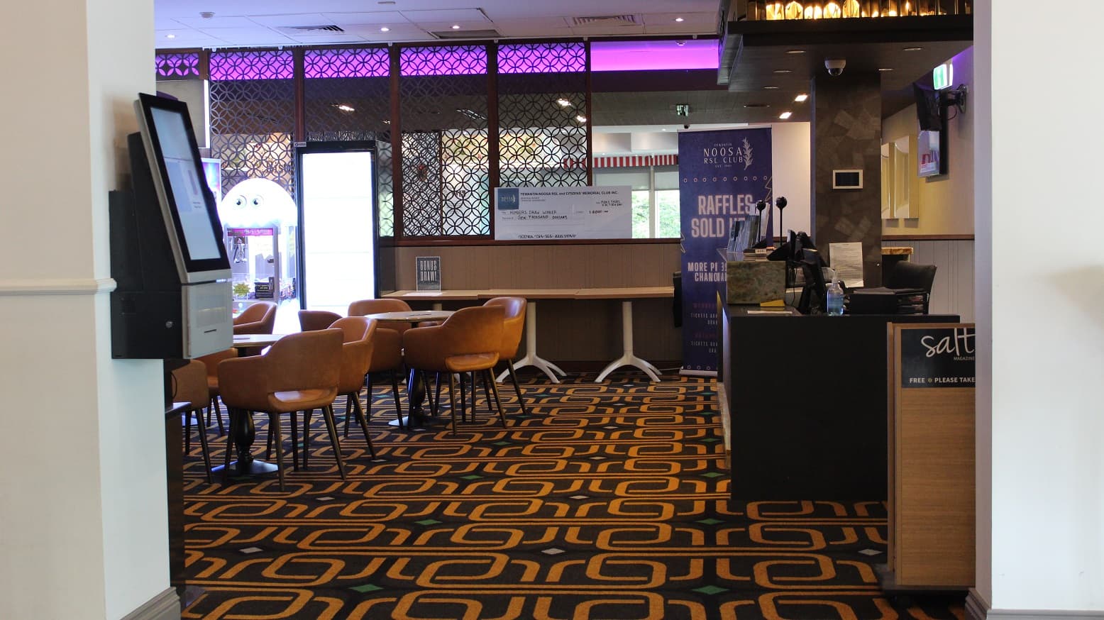 A seating space with brown chairs arranged around tables on a military-themed rug. A check-in device is on the right pillar. A demo cheque is set on the cubicle at the end with a brown barricade, which also has three tables in front by the Noosa raffle banner. A black checking desk has on it, two screens, books, sanitizer, and other materials, and a salt magazine box on the side. Behind the desk is a chair by a beige colour board with a screen.