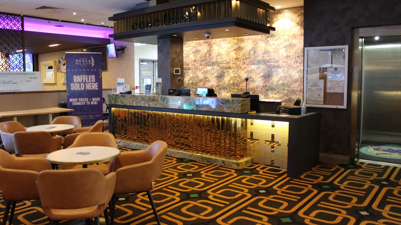Another view of the previous image focusing on the well-lit spectacular counter decked out in strong gold and army accent. It has two upright speakers, four screens, boxes, sanitizers, and booklets. An emergency exit door with an infographic is on the left and the counter looks out onto the living room, which is furnished with two sets of four brown chairs arranged around a table. as well as a Noosa RSL club banner on the side.