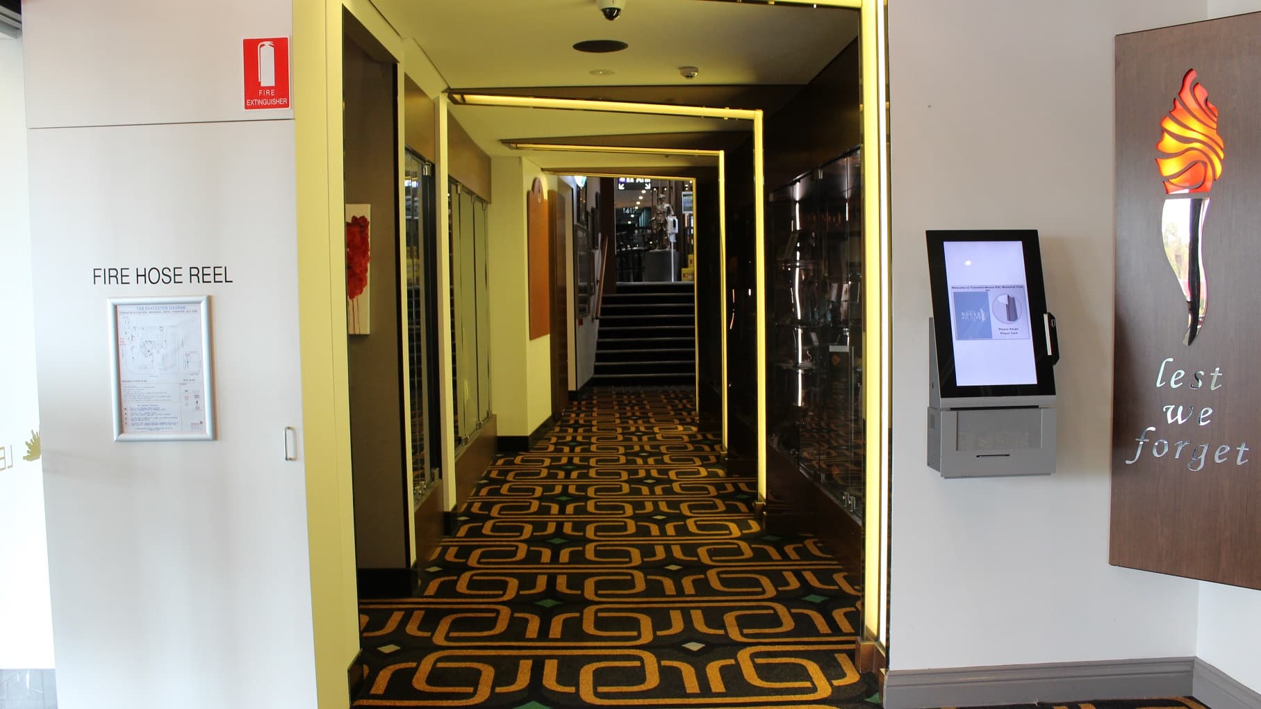 A lemon coloured-lit hallway leads to a grey-tiled step entryway with a check-in machine on the right. The artwork and glass patterns span both sides of the wall. A brown artwork, a fire hose reel, and an infographic are on the left wall. At the far end, is a sculpted soldier. The floor is covered in brown and gold.