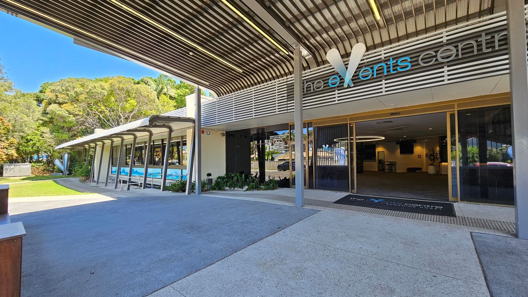 Entrance to a modern events centre with a large overhanging roof and glass doors. The sign above reads "the events centre" with stylised lettering. On the left, a pathway leads past a building with large windows and a mural, surrounded by lush greenery and trees. The ground is paved with light-coloured concrete, and the ceiling features a metal slatted design. Inside, a circular light fixture is visible.