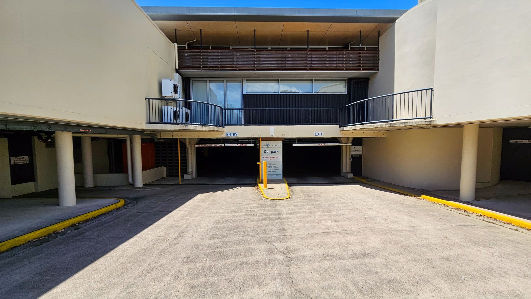 On the left, a curved road with parked vehicles and a yellow line along the edge. A large tree is visible near the centre. On the right, a building with a flat roof and a balcony, featuring air conditioning units. Below, a car park with a sign indicating "Café Open" and another sign for event parking. A small orange cone is on the pavement near the road. The ground is a mix of concrete and grass.