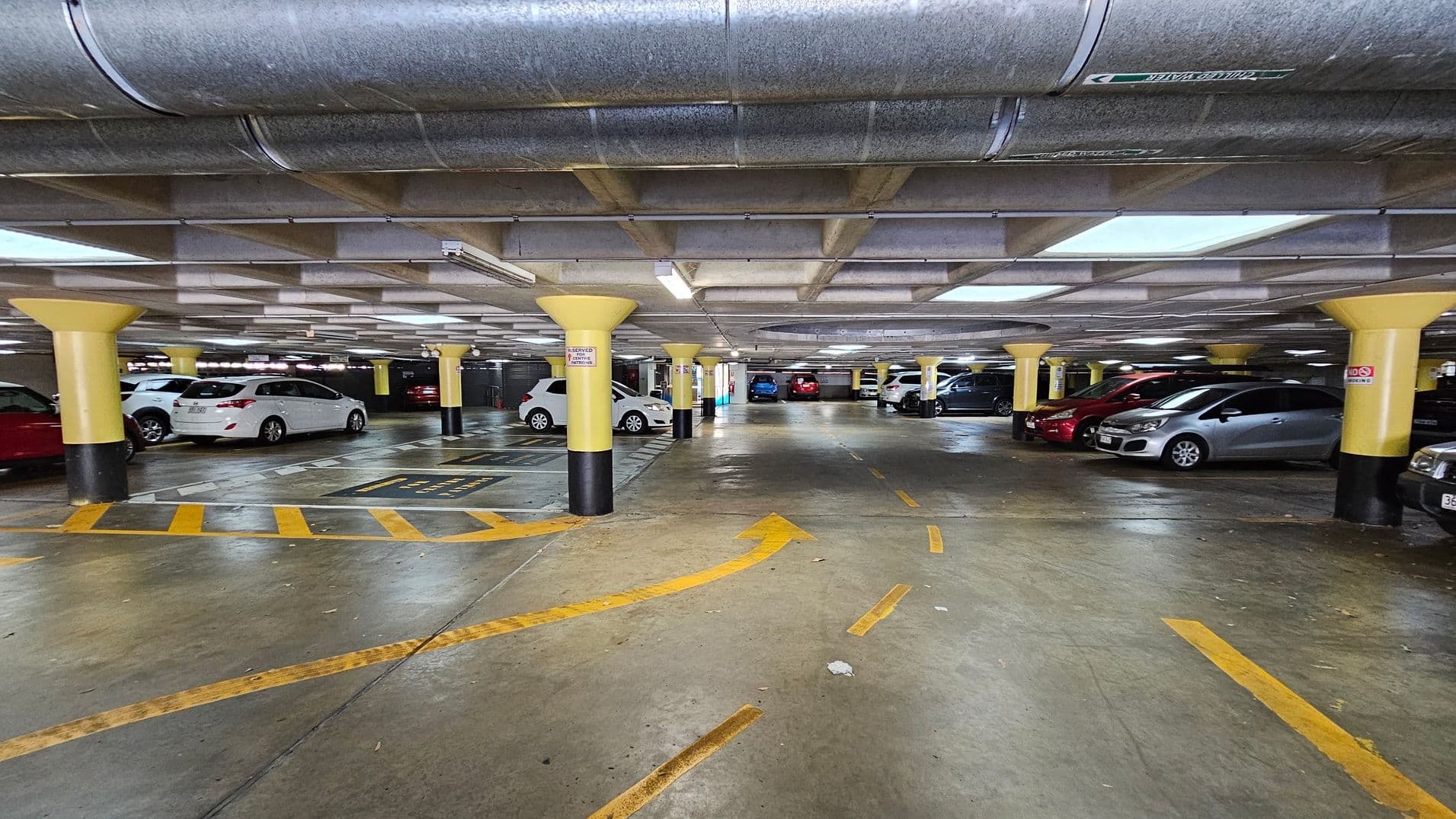 Underground car park with a concrete floor marked with yellow directional arrows and lines. Yellow and black pillars support the low ceiling, which has exposed ductwork and fluorescent lighting. Cars are parked on both sides, including white, red, and silver vehicles. The area is well-lit, and the space appears clean and organised.