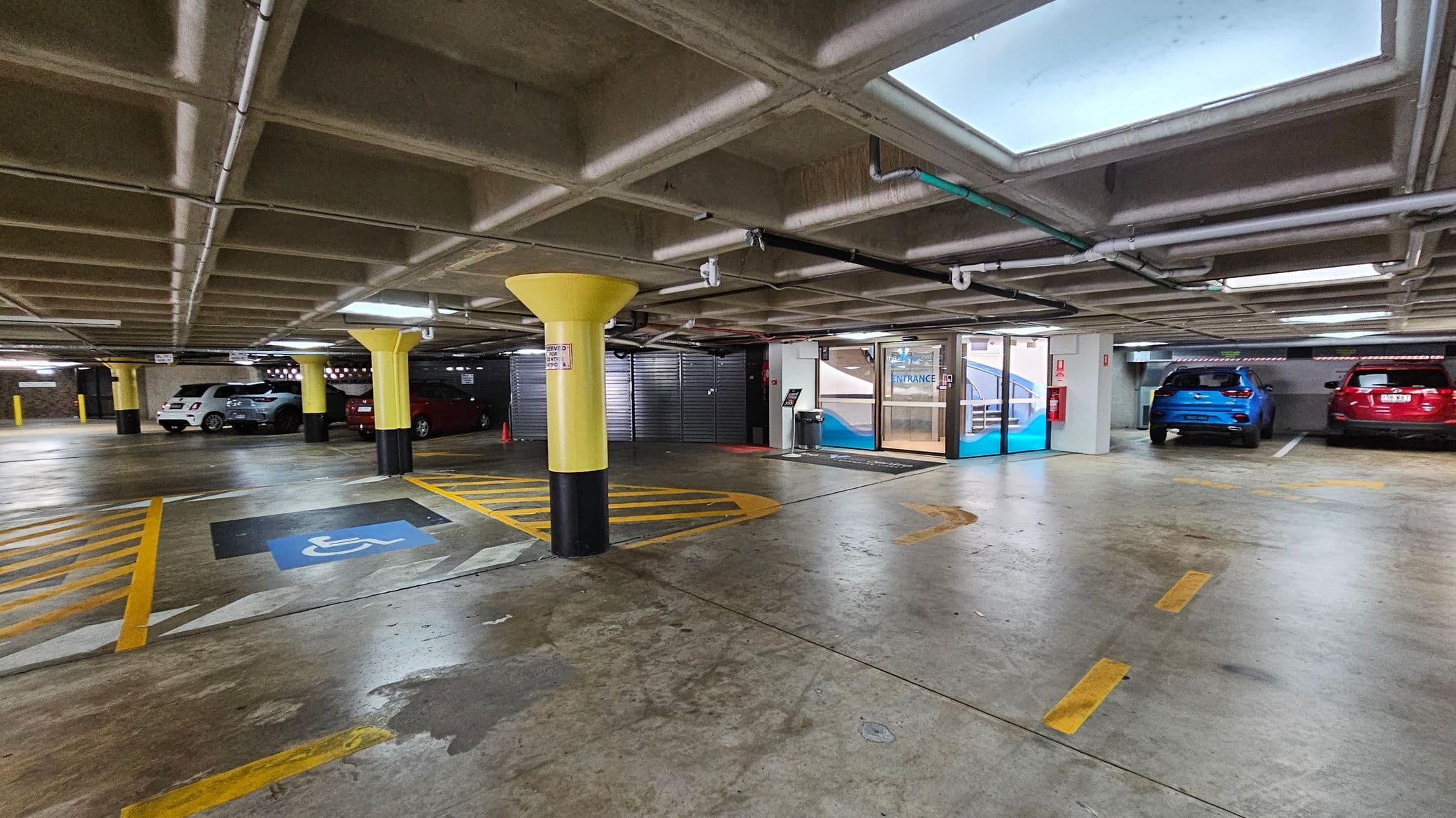 Underground car park with a concrete floor and ceiling. On the left, several cars are parked, including a white and a silver vehicle. In the centre, a yellow column stands near a marked disabled parking space with blue and yellow lines. On the right, a blue and a red car are parked. The background features a glass entrance door with a sign above it. The ceiling has visible pipes and rectangular skylights.