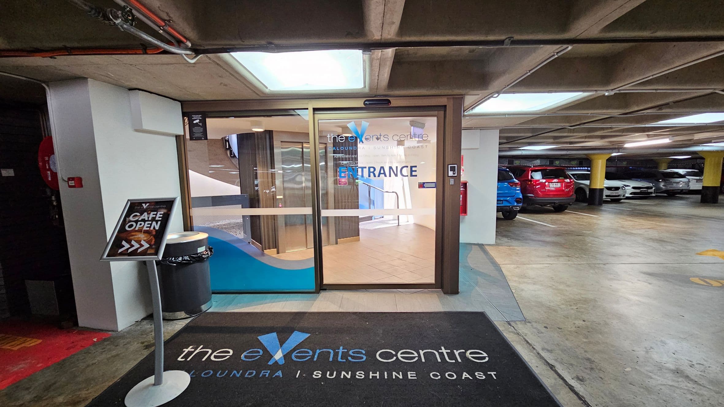 Entrance to a venue from a car park. On the left, a sign reads "CAFE OPEN" with an arrow, next to a bin. The glass door in the centre has "the events centre" and "ENTRANCE" written on it. Inside, a staircase is visible. On the right, several parked cars, including a red and a blue one, are in the car park. The floor is concrete, and the ceiling has exposed pipes and lights. A black mat with "the events centre" is on the floor in front of the door.