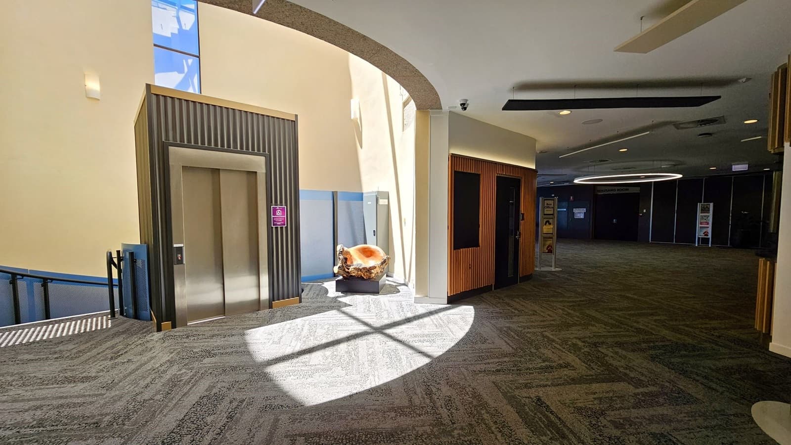 A modern interior with a lift on the left, featuring a metallic door and a striped panel design. To the right, a wooden panelled wall with a dark doorway. In the centre, a large, polished wooden sculpture sits on a pedestal, illuminated by natural light from above. The floor is carpeted in a green and grey pattern, and the ceiling has recessed lighting and a circular light fixture in the background.