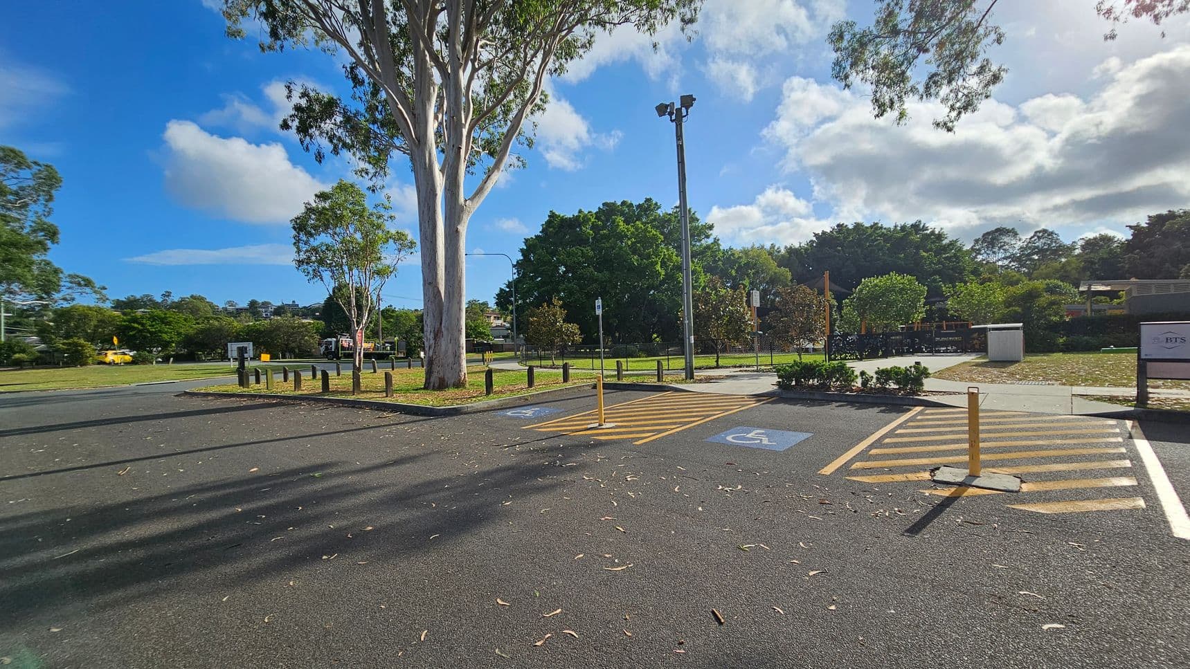 A car park with two marked disabled parking spaces in the foreground, featuring blue wheelchair symbols. A large tree stands to the left, casting shadows on the asphalt. In the background, there are more trees and a grassy area, with a fence and a small structure visible. A tall light pole is near the centre, and a sign is on the right. The ground is scattered with leaves and debris.