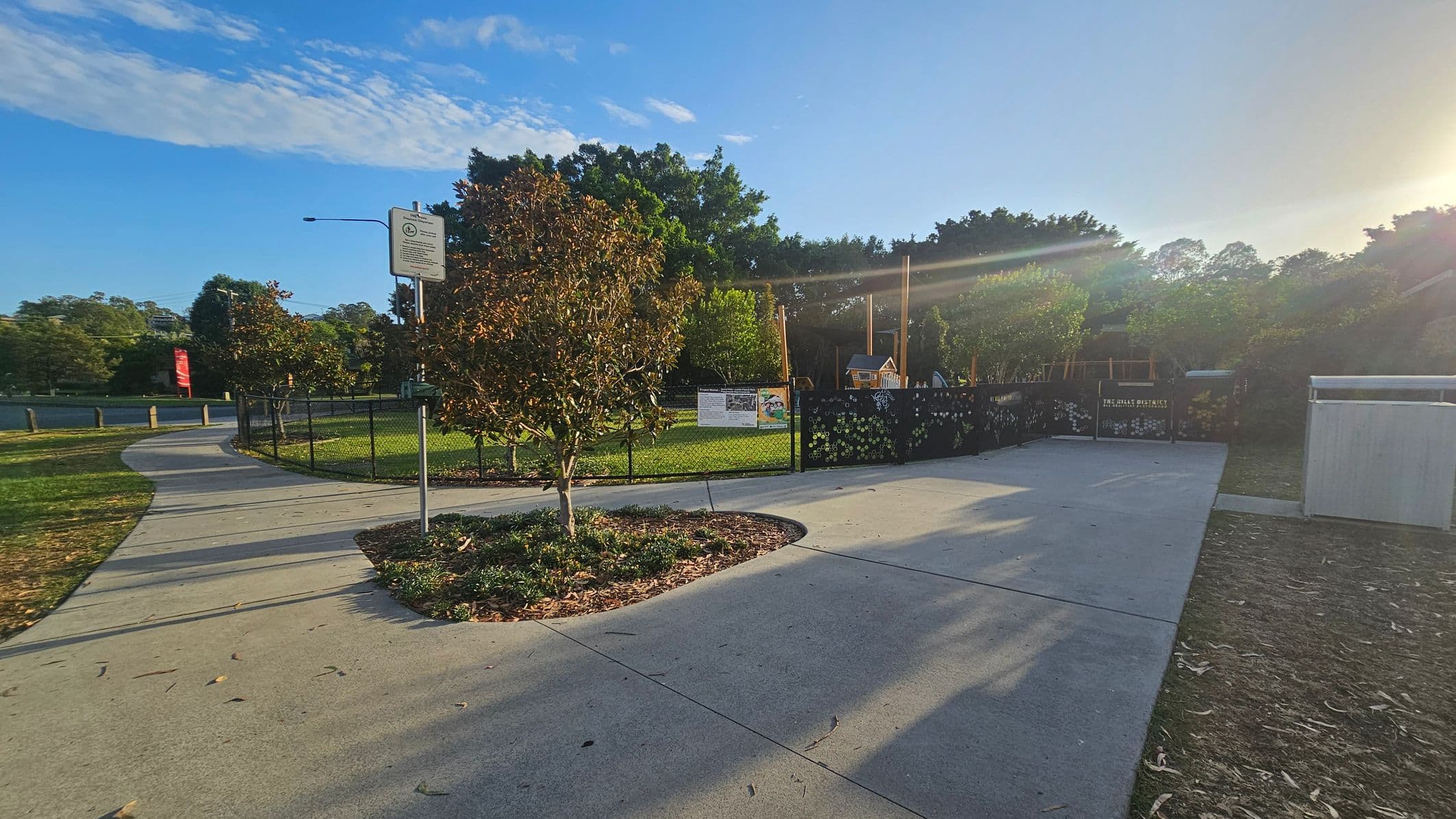A paved pathway curves to the left, bordered by grass and small trees. A signpost stands near the path. To the right, a fenced area encloses a playground with a small playhouse and equipment. The fence has decorative elements and a sign. Trees and greenery fill the background, with sunlight streaming from the right, casting long shadows on the ground. The sky is clear with some clouds.
