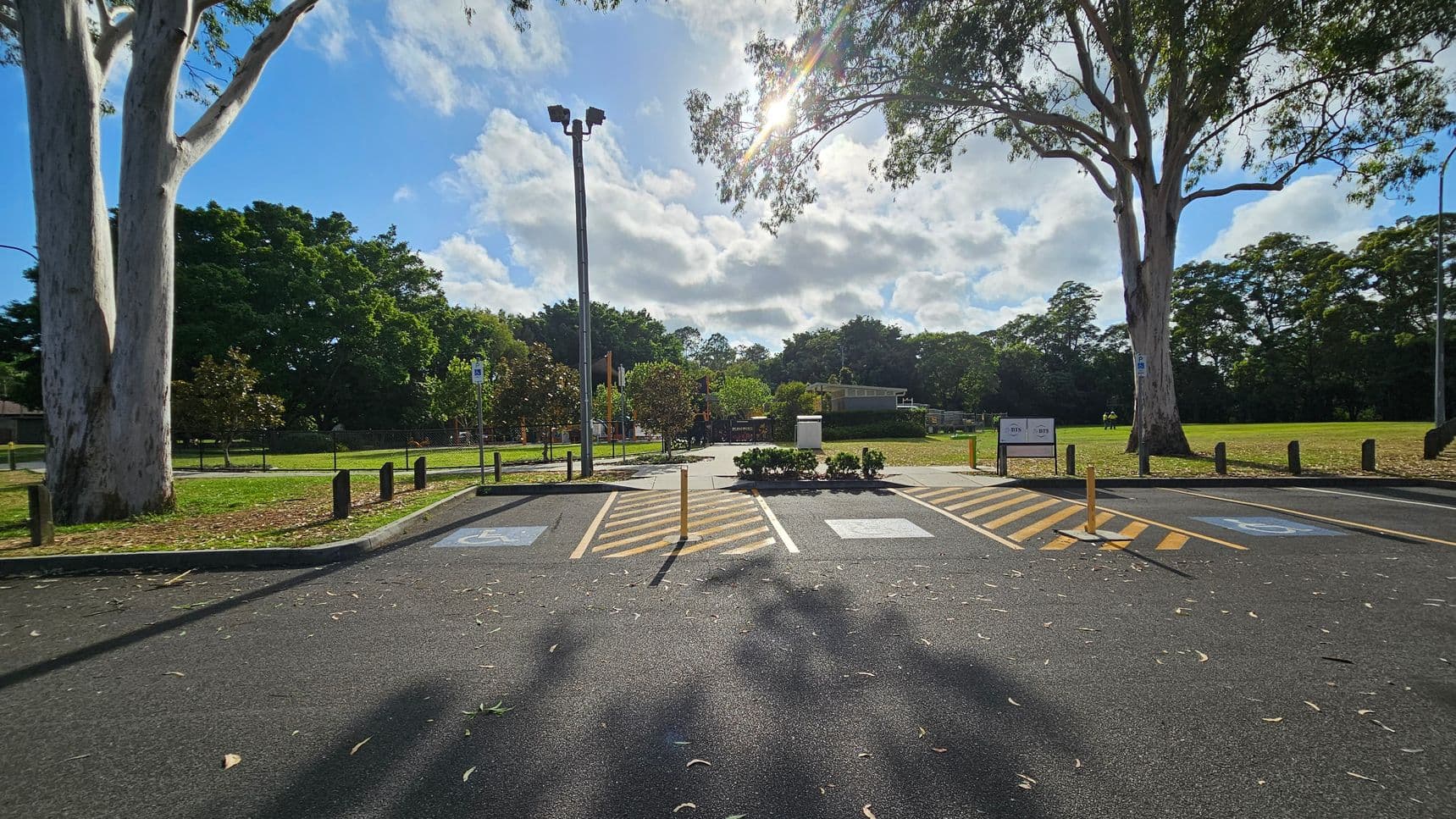 A car park with designated disabled parking spaces marked in blue and yellow lines in the foreground. Two large trees stand on either side, casting shadows on the asphalt. In the background, a grassy area with more trees and a playground is visible. A tall light pole is centrally located, and the sun shines through the clouds, creating a bright spot in the sky. A sign is visible near the trees on the right.