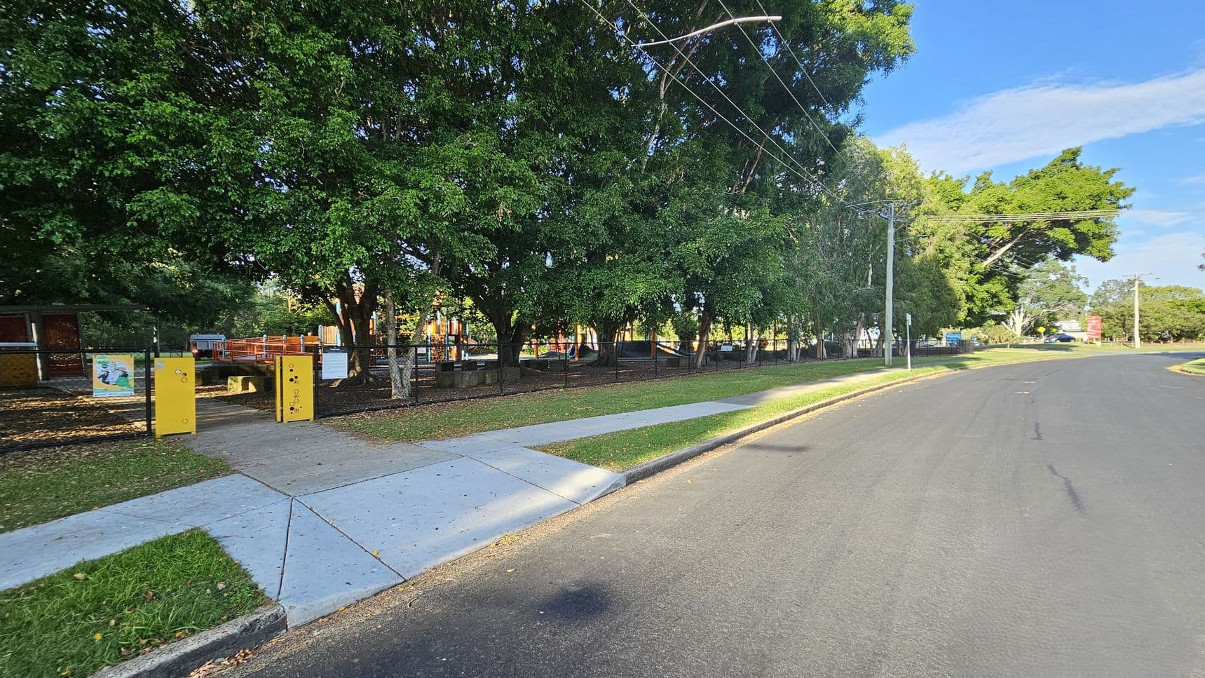 A quiet street with a wide road on the right and a footpath on the left. The left side features a park enclosed by a black fence, with large green trees providing shade. Yellow entry gates and signs are visible at the park entrance. The park contains playground equipment in the background. Power lines run overhead, and a grassy verge separates the footpath from the road.