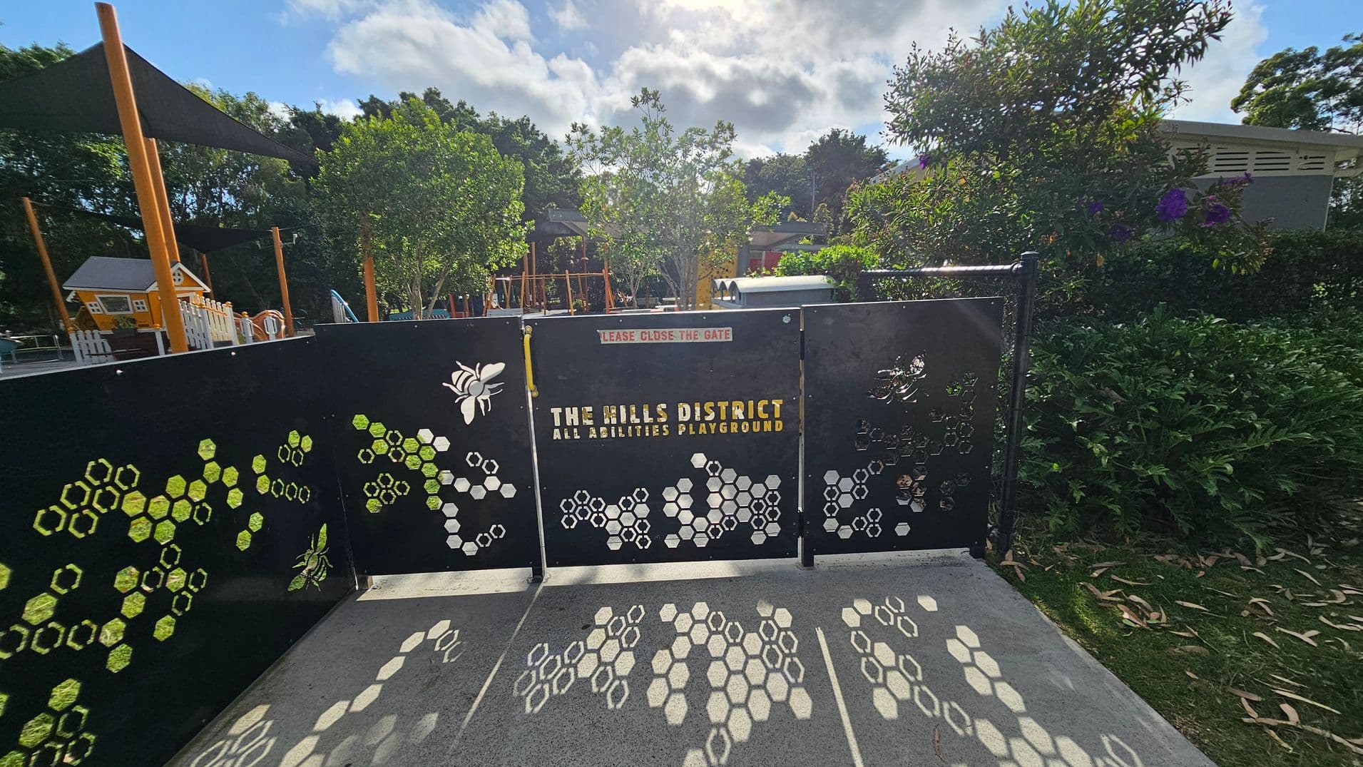 A black gate with honeycomb and bee designs, labelled "The Hills District All Abilities Playground," stands in the foreground. The ground is concrete with honeycomb patterns. Behind the gate, there are trees and playground equipment, including a small house structure and slides. The background is filled with lush greenery and a partly cloudy sky.