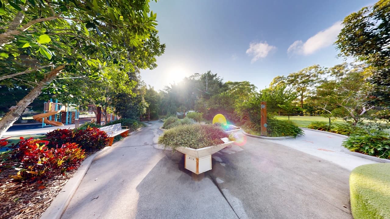 A garden scene with large, hexagonal planters filled with various plants and flowers on the left. In the centre, a pathway leads through the garden, with colourful, dome-shaped structures resembling beehives on the right. The background is filled with lush green trees and a glimpse of a playground with a slide. The ground is paved, and the area is well-maintained, with vibrant greenery throughout.