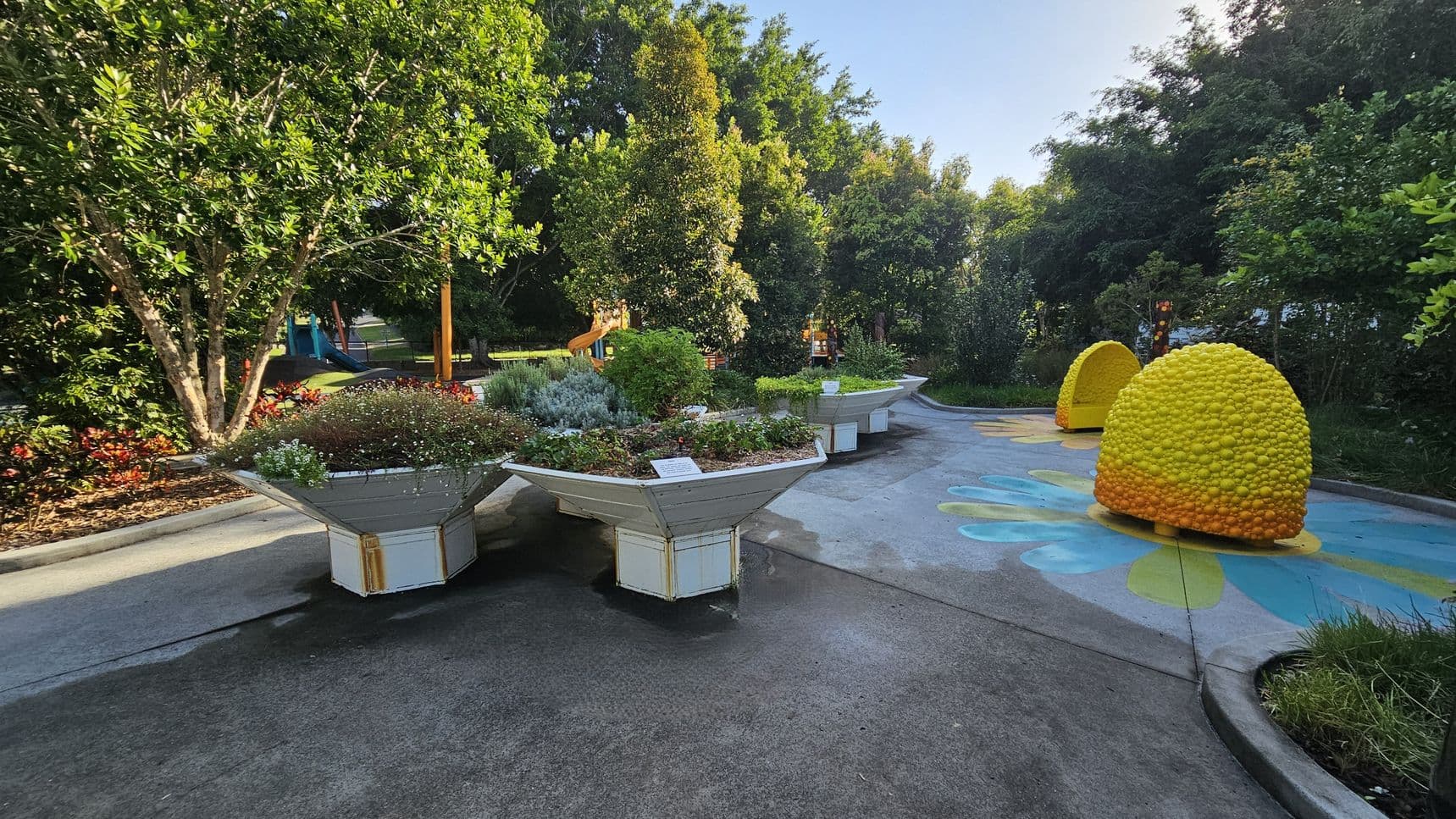 A garden scene with large, hexagonal planters filled with various plants and flowers on the left. In the centre, a pathway leads through the garden, with colourful, dome-shaped structures resembling beehives on the right. The background is filled with lush green trees and a glimpse of a playground with a slide. The ground is paved, and the area is well-maintained, with vibrant greenery throughout.