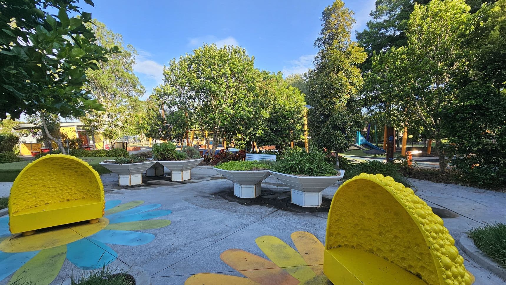A vibrant park scene with two large, yellow, semi-circular benches resembling lemon slices on the left and right. The ground features colourful flower patterns. In the centre, there are several large, white, hexagonal planters filled with greenery. The background is filled with lush trees and a playground area with slides and climbing structures. The sky is clear and blue, and the area is well-lit, suggesting a sunny day.