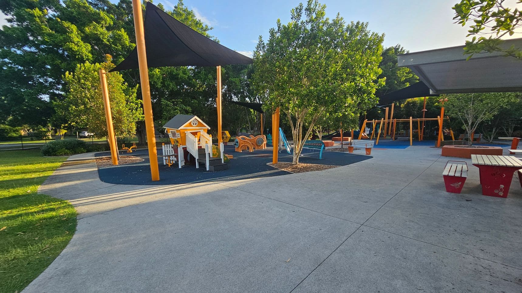 A playground with a small playhouse and various play equipment on the left, surrounded by trees and shaded by large triangular canopies. The ground is covered with a mix of soft play surfaces and concrete paths. On the right, there are picnic tables with red benches. The background features more trees and additional play structures, including swings. The area is well-maintained and spacious, with a mix of sun and shade.