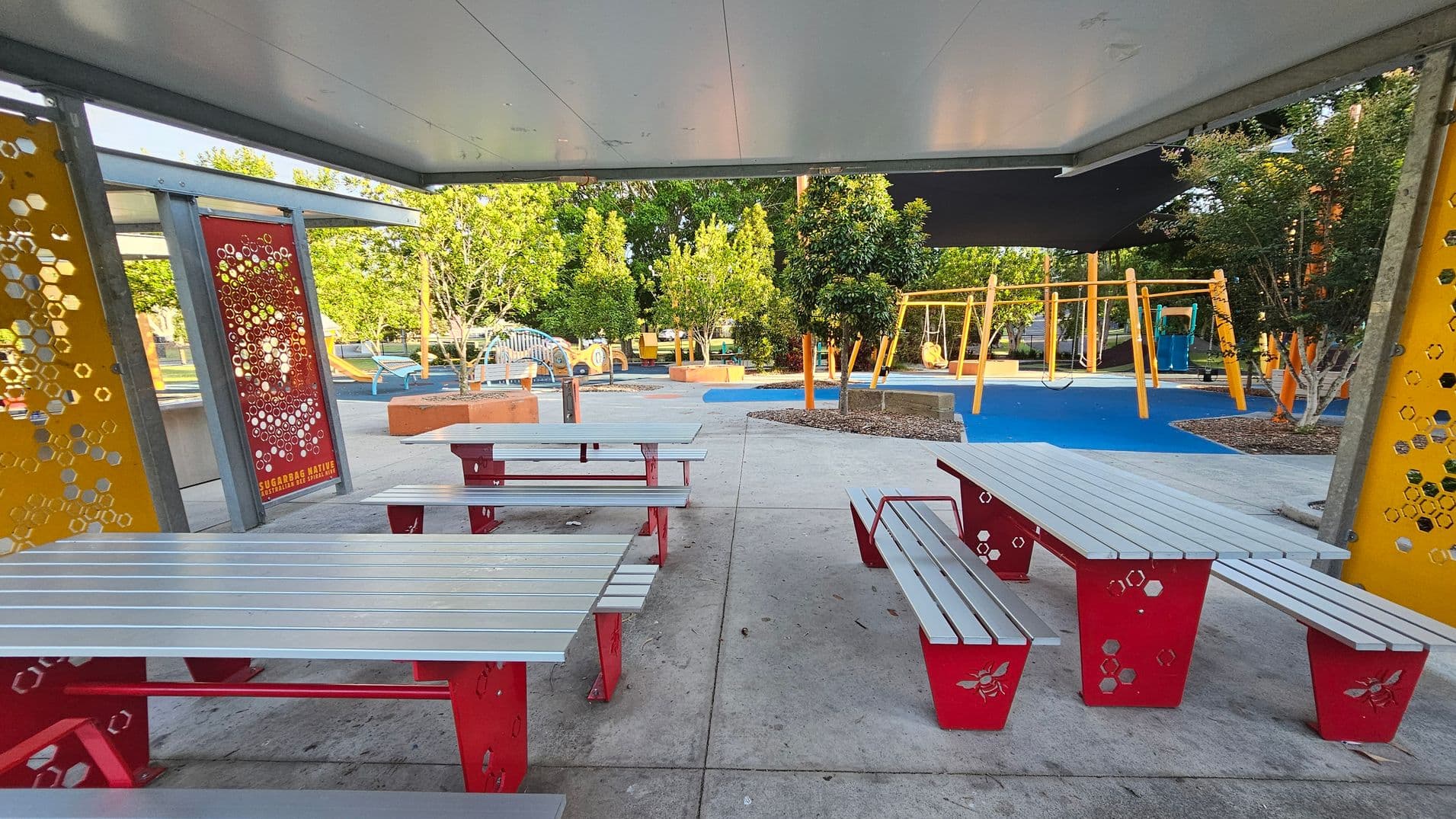 Under a grey ceiling, there are several picnic tables with red metal legs and grey tops. The floor is concrete. In the background, a playground features swings and climbing structures on a blue rubber surface. Trees and shrubs are scattered throughout the area. Decorative panels with hexagonal patterns and bee motifs are on the left and right, with one labelled "Sugarbag Native Bee."