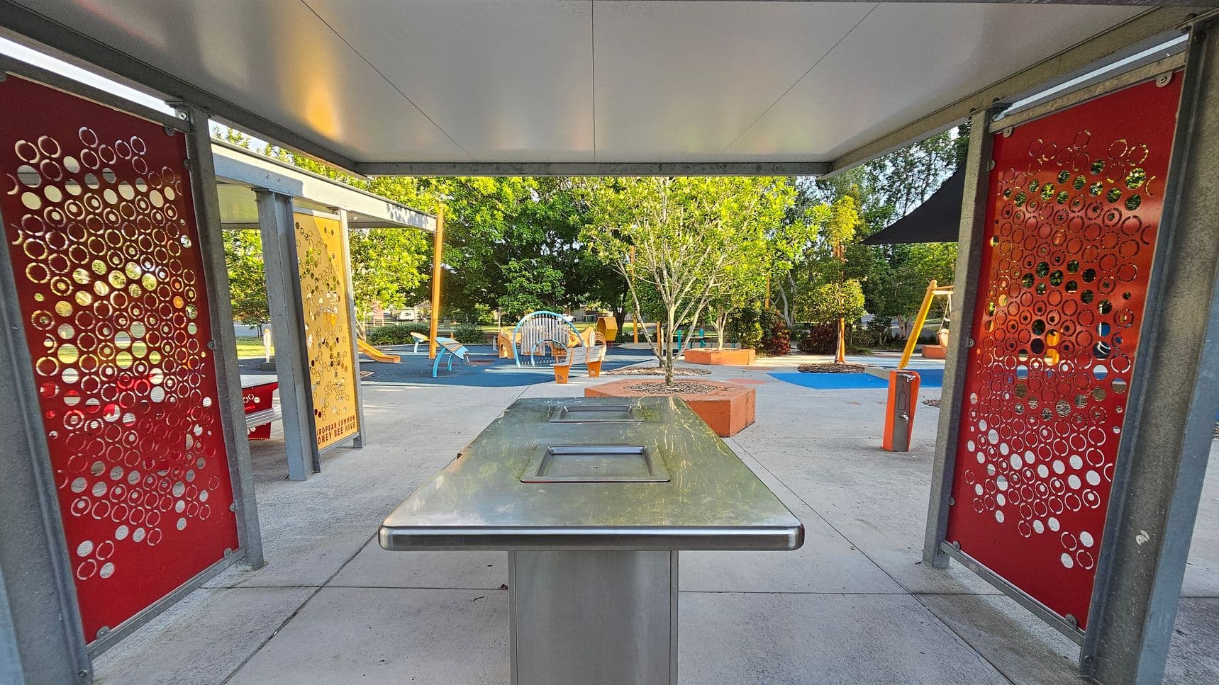 A covered outdoor area with a stainless steel table in the centre. On the left and right are red panels with circular cut-out patterns. In the background, a playground with various equipment, including a slide and climbing structures, is visible. Trees and shrubs are scattered throughout, with some planted in orange-bordered planters. The ground is a mix of concrete and blue rubberised surfaces. The ceiling is flat and grey.