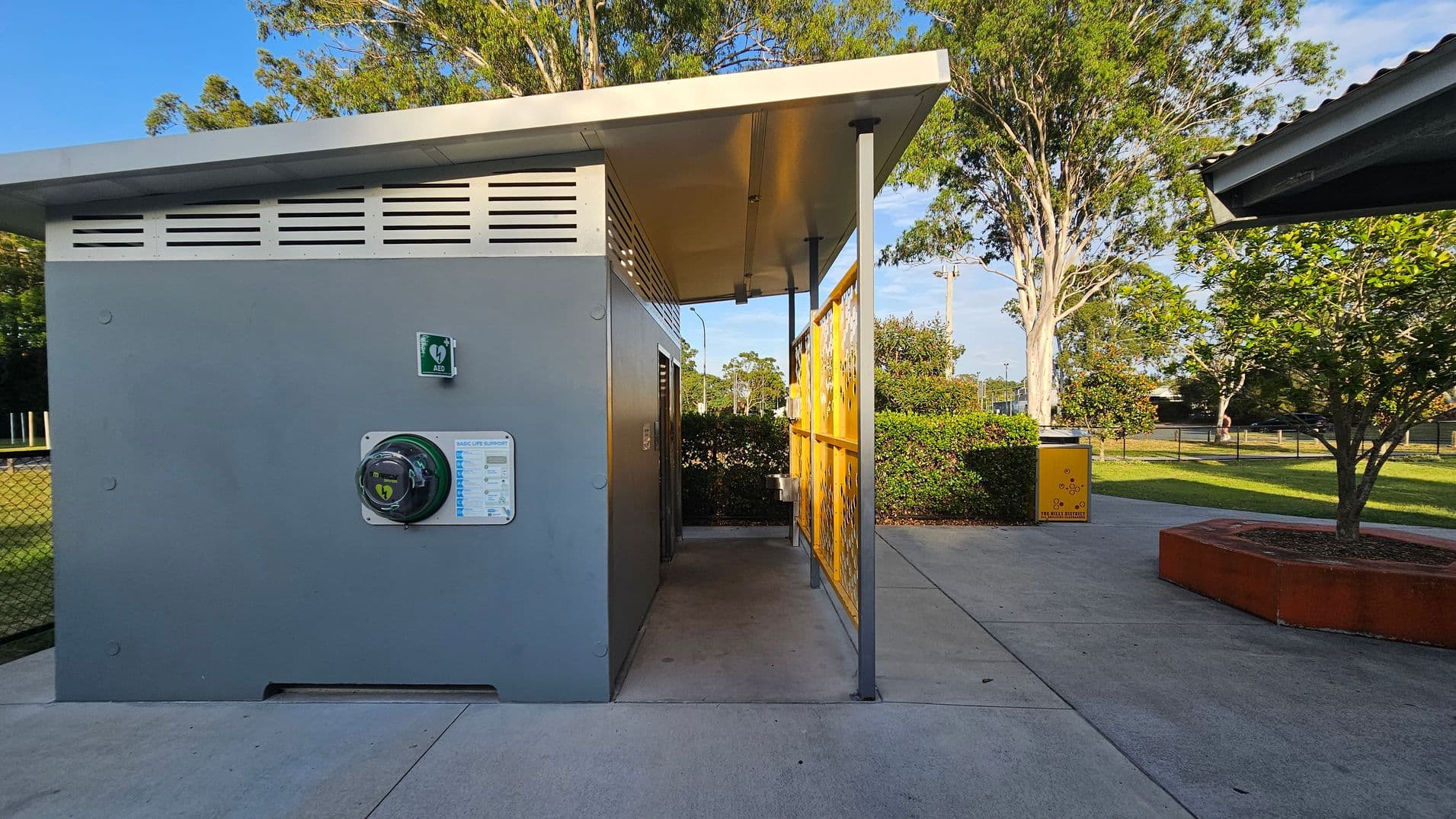A small grey building with a slanted roof is on the left, featuring an automated external defibrillator (AED) mounted on the wall. A yellow decorative panel is on the right side of the building. In the background, there are trees and a grassy area with a yellow bin. The ground is paved with concrete, and a tree is planted in a red circular planter on the right.