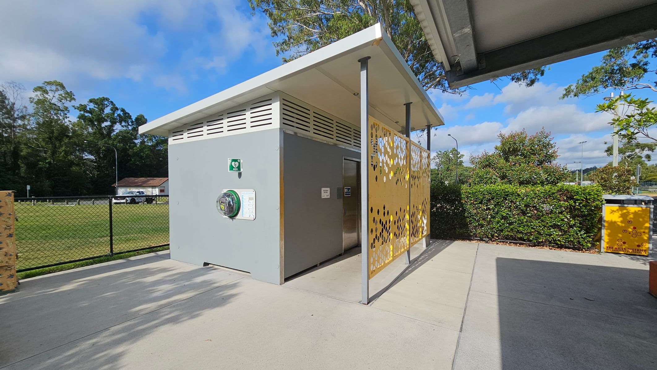 A small, modern public toilet structure with a grey exterior and a slanted white roof. On the right, there's a decorative yellow panel with circular cutouts. A defibrillator is mounted on the wall. In the background, a grassy field is enclosed by a black chain-link fence, with a parked white vehicle and a small building visible. Bushes and trees are scattered around, and a yellow bin is on the right. The ground is paved with concrete.