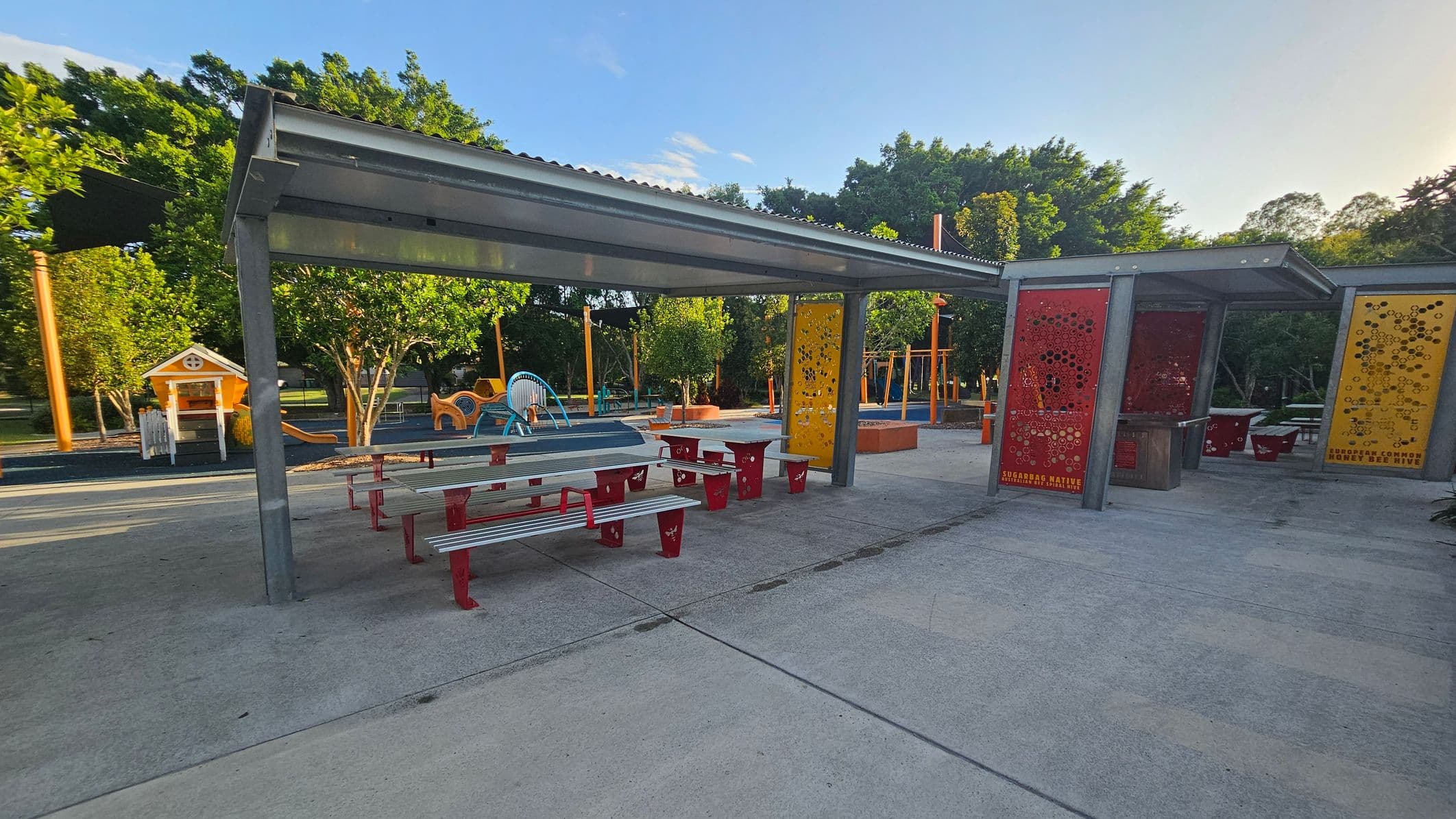 A park area with a covered seating section featuring metal picnic tables with red legs and benches. The shelter has a corrugated metal roof. To the right, there are decorative panels in red and yellow with circular cutouts. In the background, a playground with various equipment, including a small playhouse and climbing structures, is visible. The area is surrounded by trees, and the ground is paved with concrete.