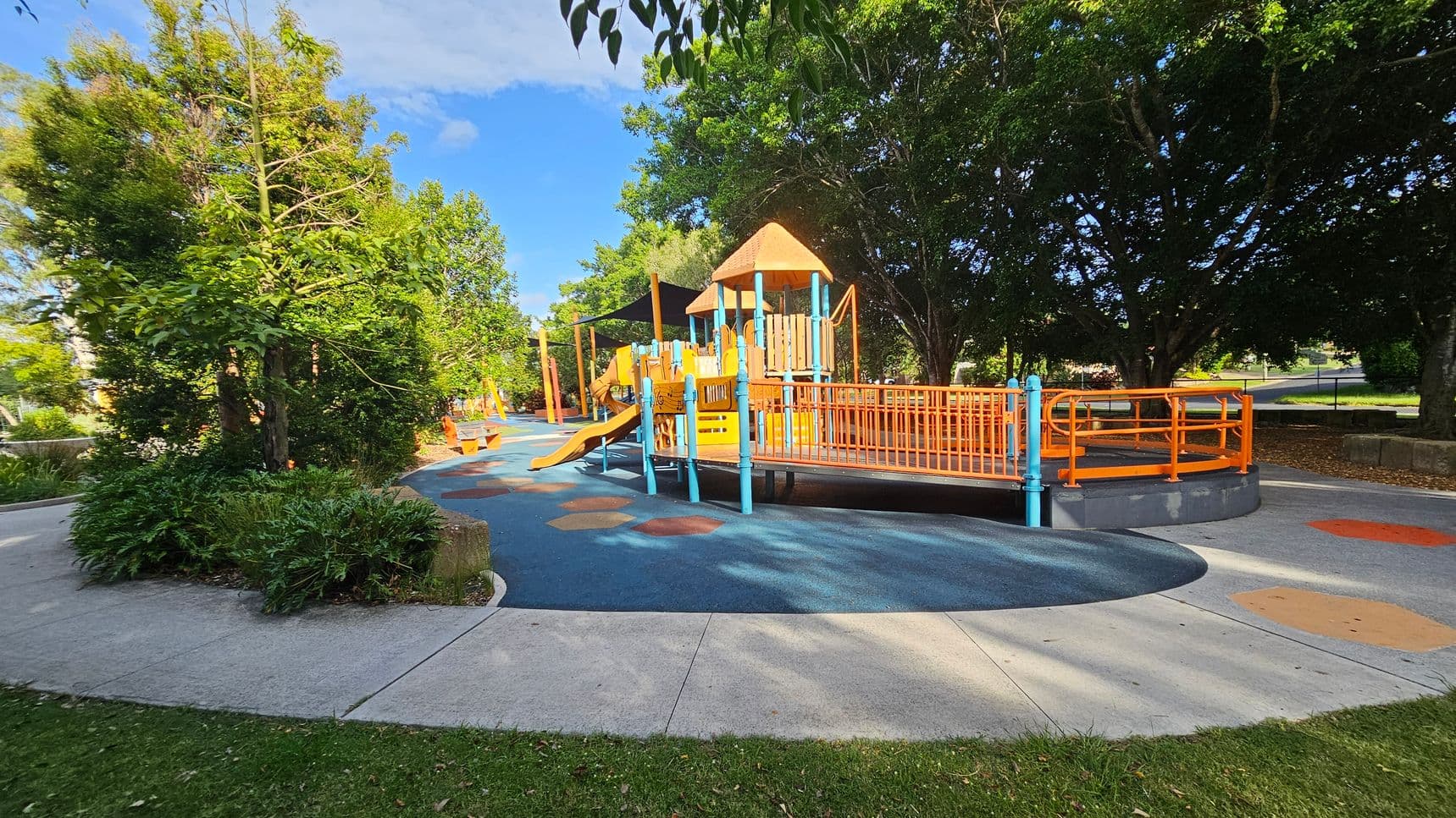 A playground with a colourful play structure featuring orange and blue elements, including slides and ramps, is in the centre. The ground is covered with a blue and orange rubber surface. On the left, there are lush green trees and shrubs. In the background, more trees provide shade. The foreground has a concrete path and grass.