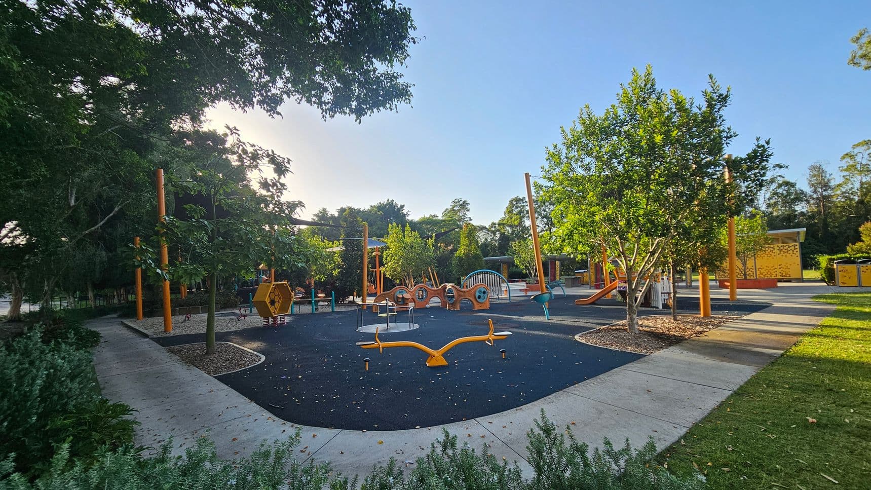 A playground with various equipment is surrounded by trees and greenery. On the left, there are large trees providing shade. In the centre, a black rubber play surface features a yellow seesaw, a hexagonal climbing structure, and other play equipment. To the right, more trees and a slide are visible. The ground is a mix of concrete paths and grass, with some mulch around the trees. The background includes more trees and a yellow structure.