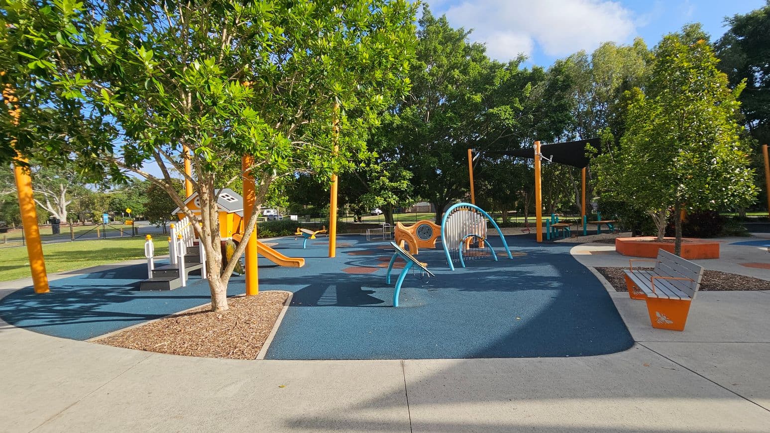 A playground with various equipment on a blue rubber surface. On the left, there's a tree with green leaves and an orange slide. In the centre, a climbing structure with blue arches and a small orange playhouse. On the right, a bench with orange accents and another tree. The background features more trees and a shaded area with a black canopy. The ground is a mix of concrete paths and mulch around the trees.