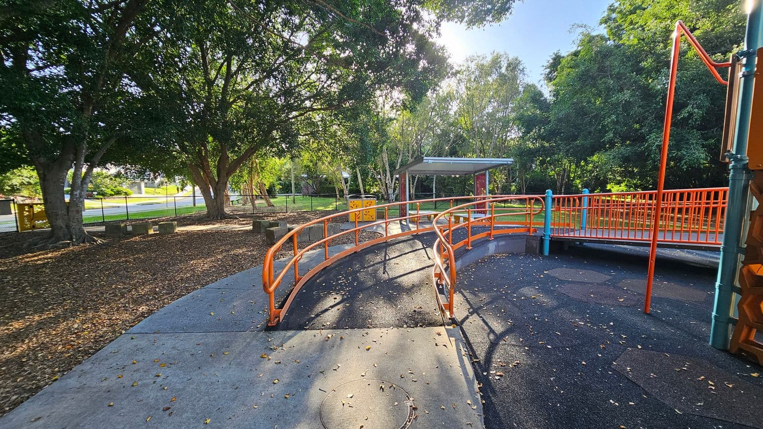 A playground with an orange railing bridge in the centre, leading to a covered area in the background. The ground is a mix of concrete and rubberised surface with scattered leaves. On the left, large trees provide shade, and a fence runs along the perimeter. On the right, more playground equipment is partially visible. The area is surrounded by greenery, creating a natural setting.