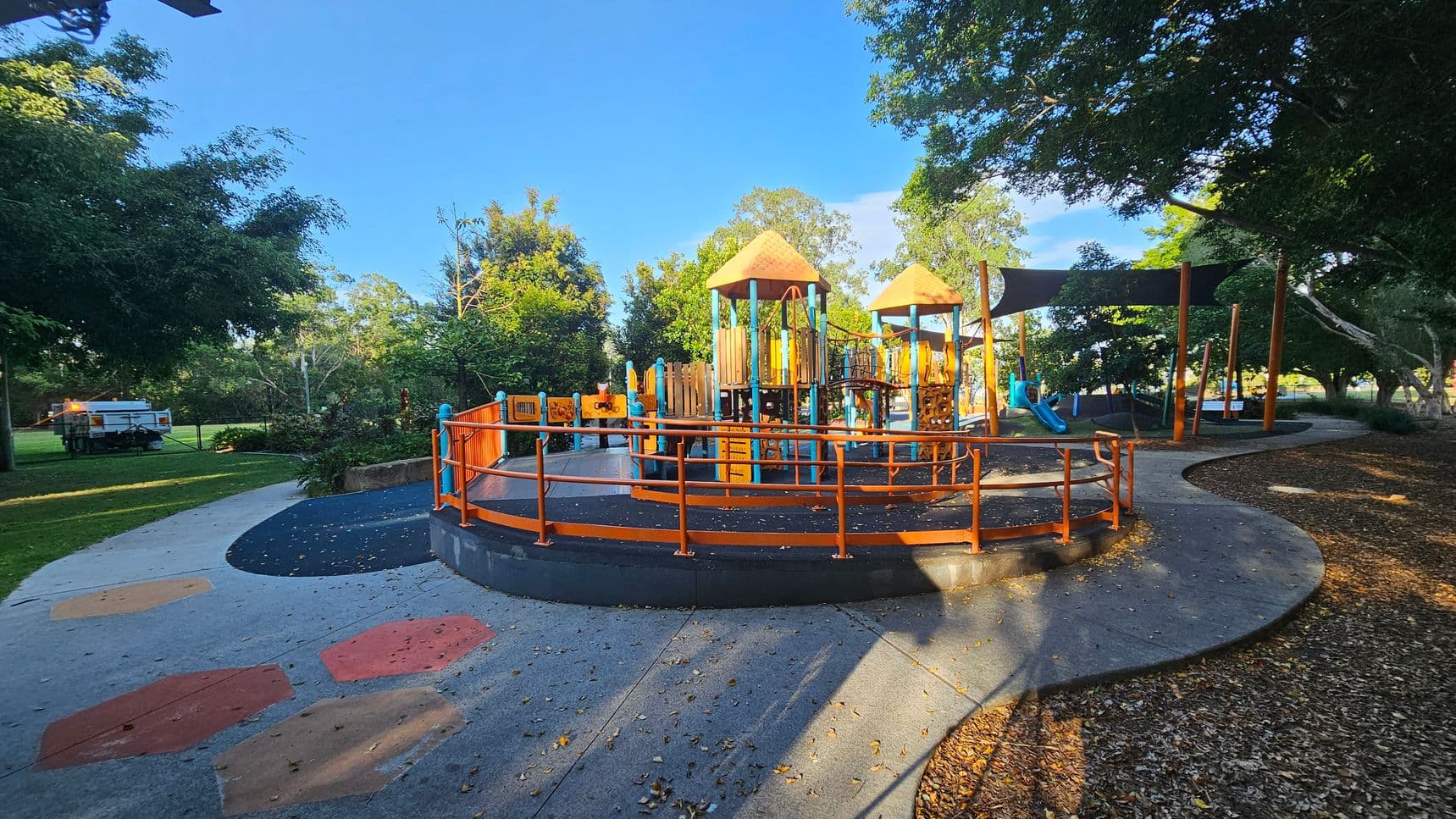 A playground with colourful equipment featuring orange and blue structures, including slides and climbing frames, is in the centre. The ground is a mix of concrete and rubber surfaces with geometric patterns. Trees surround the area, providing shade, and a path curves around the playground. A white utility vehicle is parked on the grass to the left. The background is filled with lush greenery and more trees.