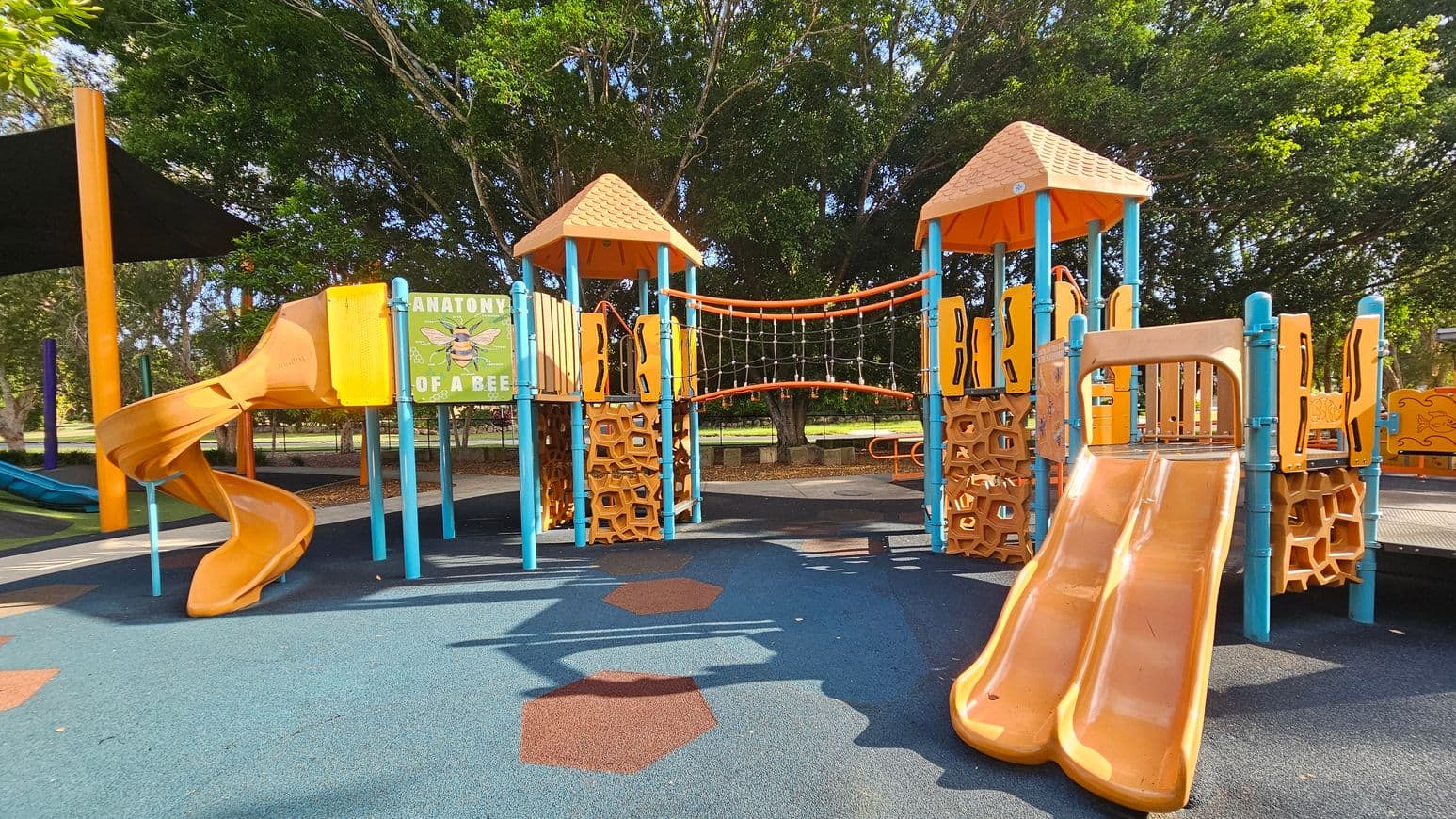A children's playground with two orange slides, one spiral on the left and a double slide on the right. The structures are supported by blue poles and feature orange hexagonal patterns. A sign titled "Anatomy of a Bee" is visible on the left. An orange rope bridge connects two towers with orange roofs. The ground is covered with blue and orange rubber tiles. Trees are in the background, providing shade.