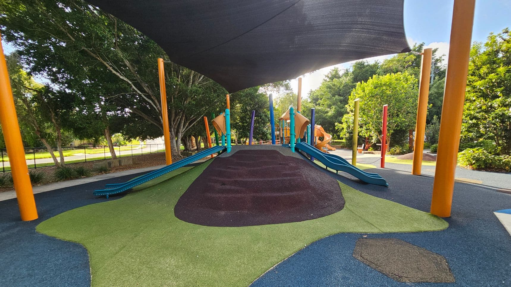 A playground with a central mound covered in green and brown rubber surfacing, flanked by blue slides. Bright orange and blue poles support a large black shade canopy overhead. In the background, there are trees and additional playground equipment, including orange slides. The ground is a mix of blue and green rubberised material, and the area is surrounded by lush greenery.