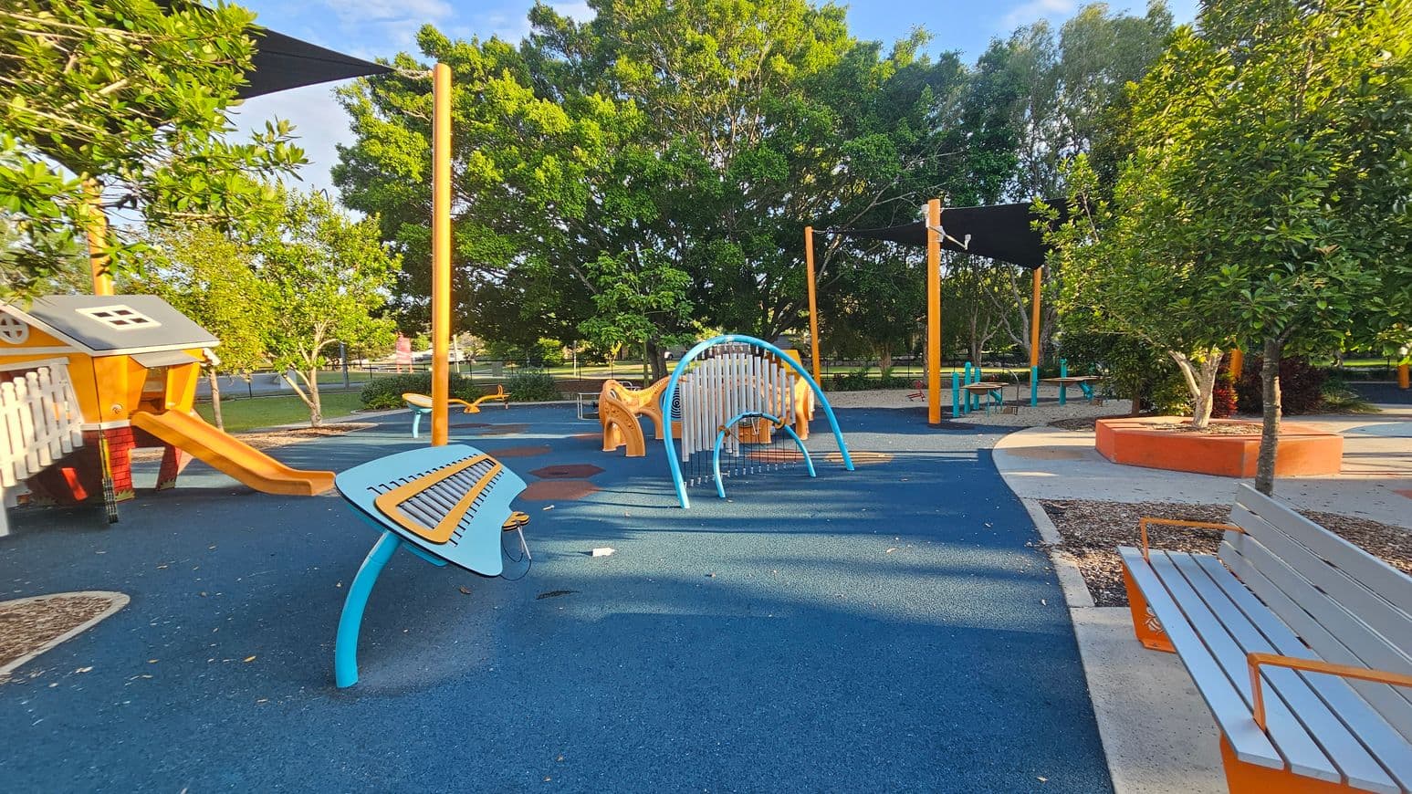 A playground with a blue rubber floor. On the left, there's a playhouse with a slide and a musical play structure. In the centre, a curved climbing frame is visible. To the right, a bench with orange supports is near a tree planted in a circular orange planter. In the background, there are more trees and a shaded area with additional seating.
